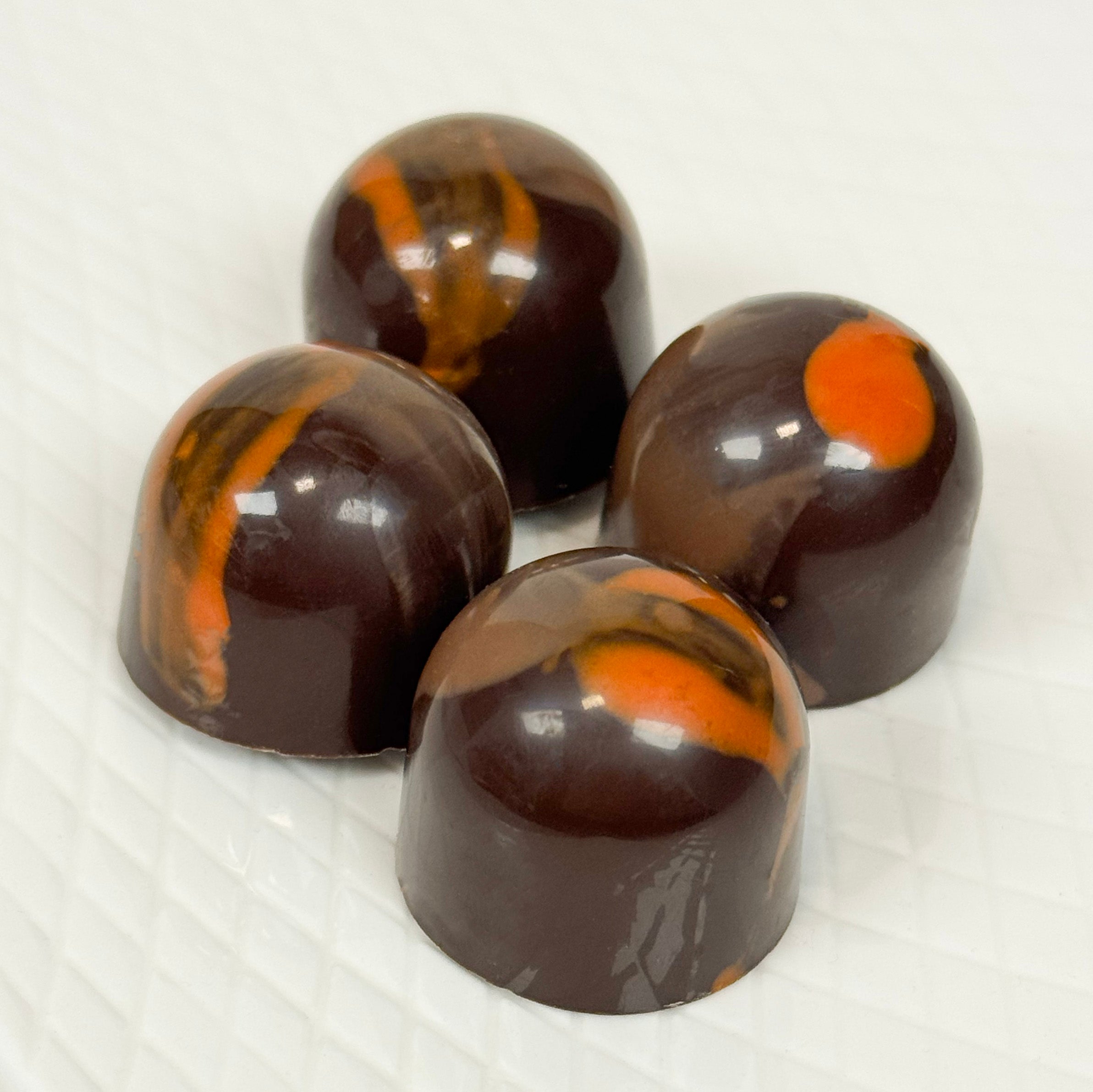 Four glossy dark chocolate domed confections with orange swirls, arranged on a white textured surface.