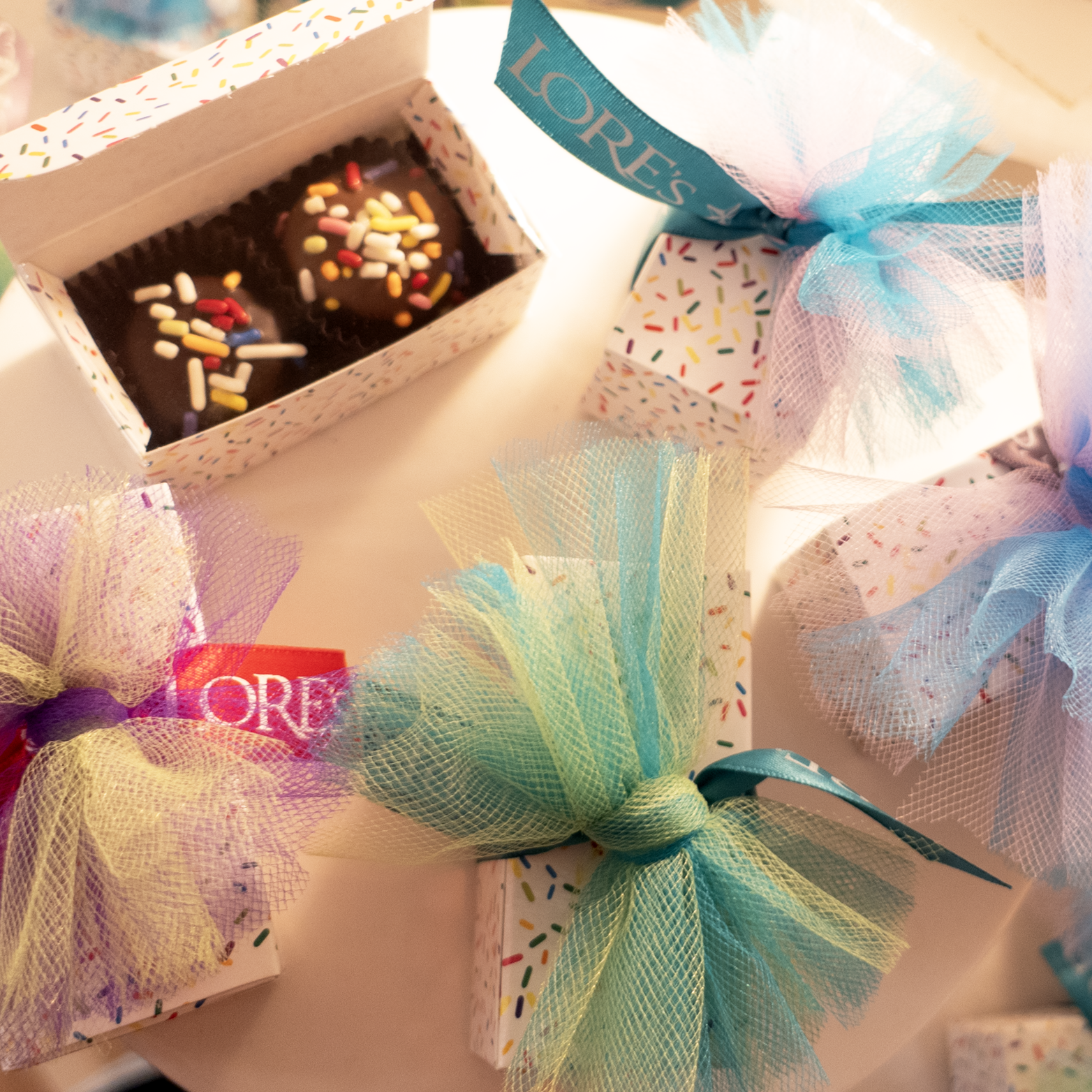 a white box open to show two Birthday Cake Cream Chocolates with s[rinkles surrounded by little boxes with rainbow sprinkles and colorful tulle ribbons.