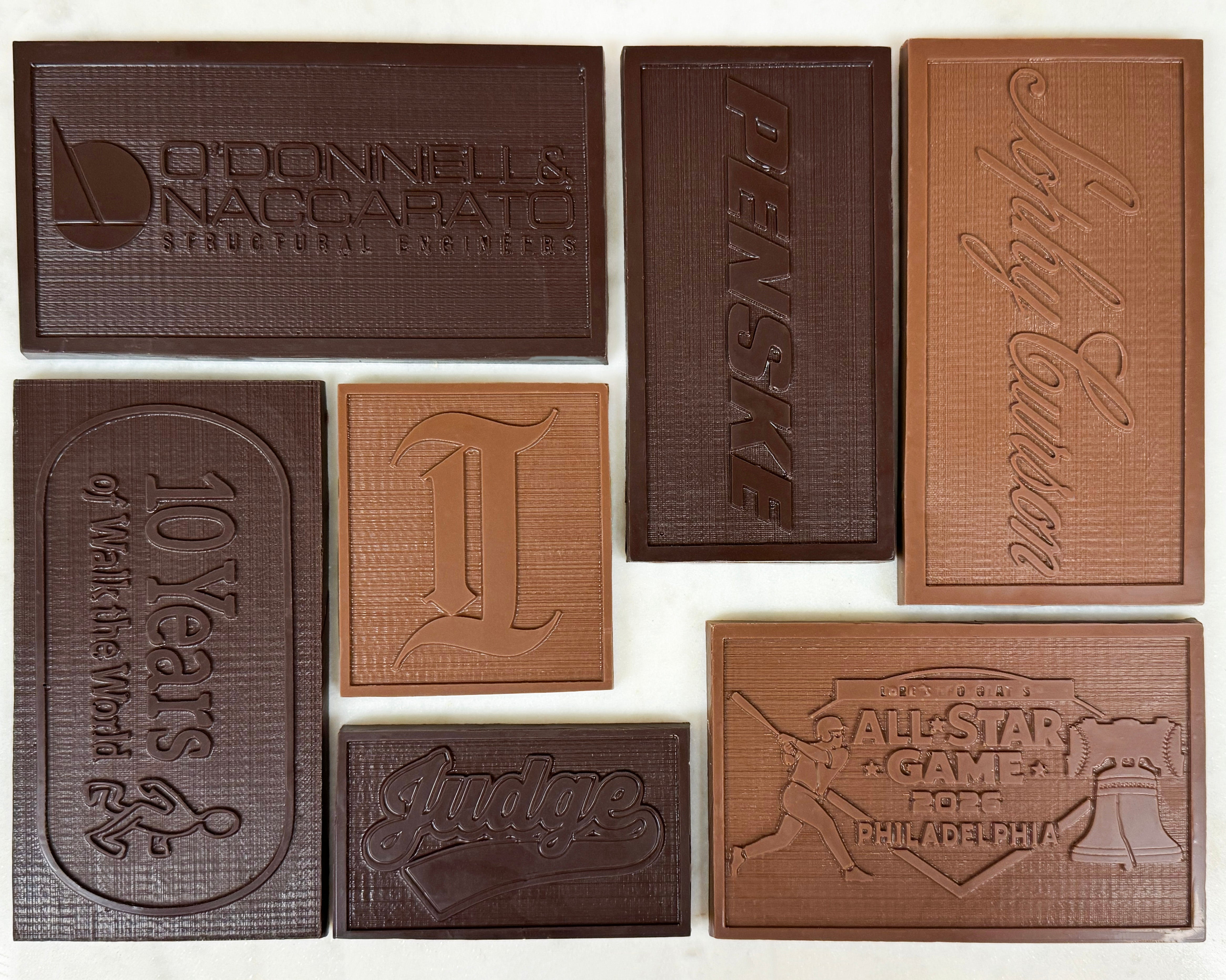 A set of seven custom embossed chocolate bars arranged on a white background, featuring various business logos such as O'Donnell's Nascarato, Penske, Jimmy Gunner, 10 Years of Make the World, Judge, and All-Star Game Philadelphia.