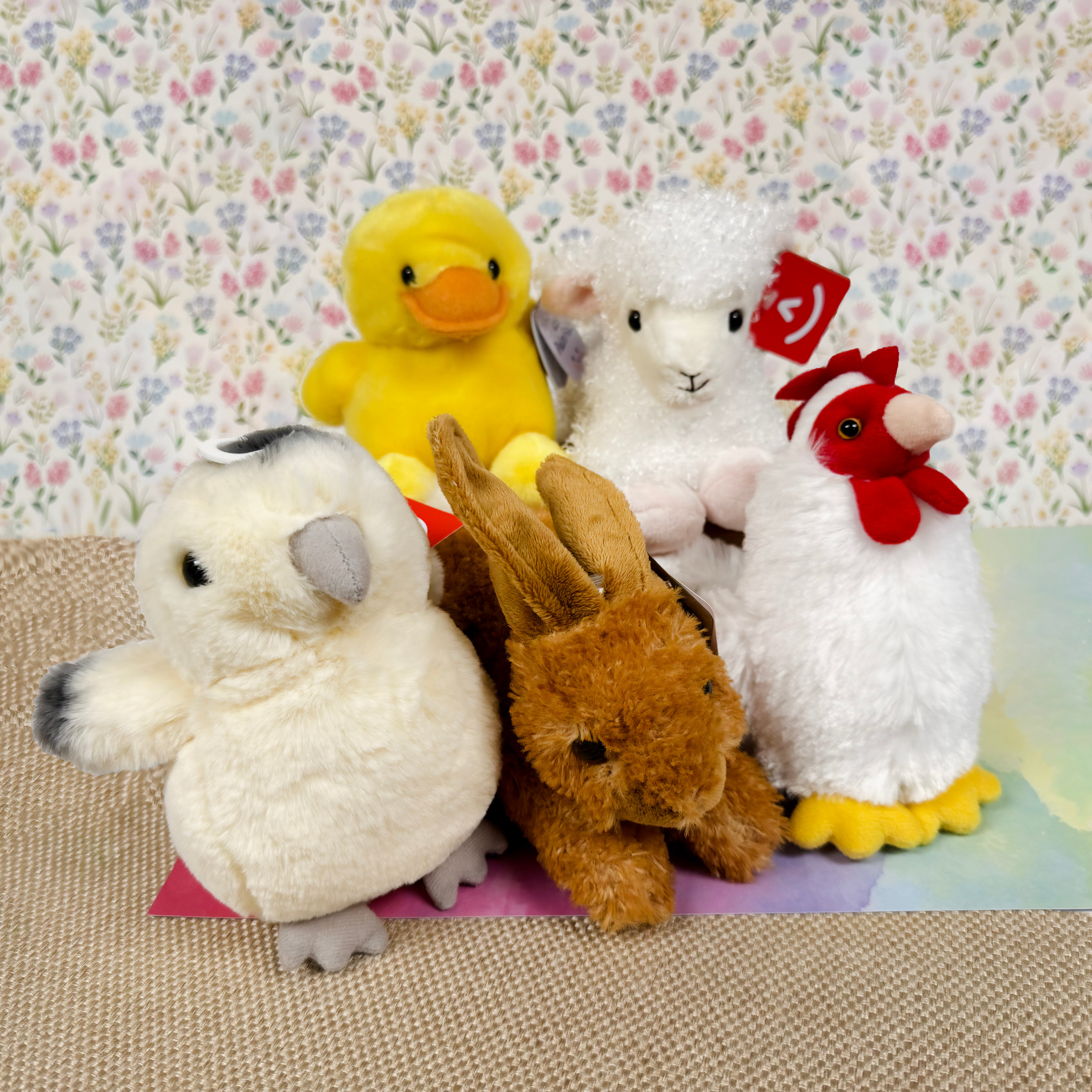 Five plush toys including a duck, sheep, chick, rabbit, and chicken, on a floral-patterned background.