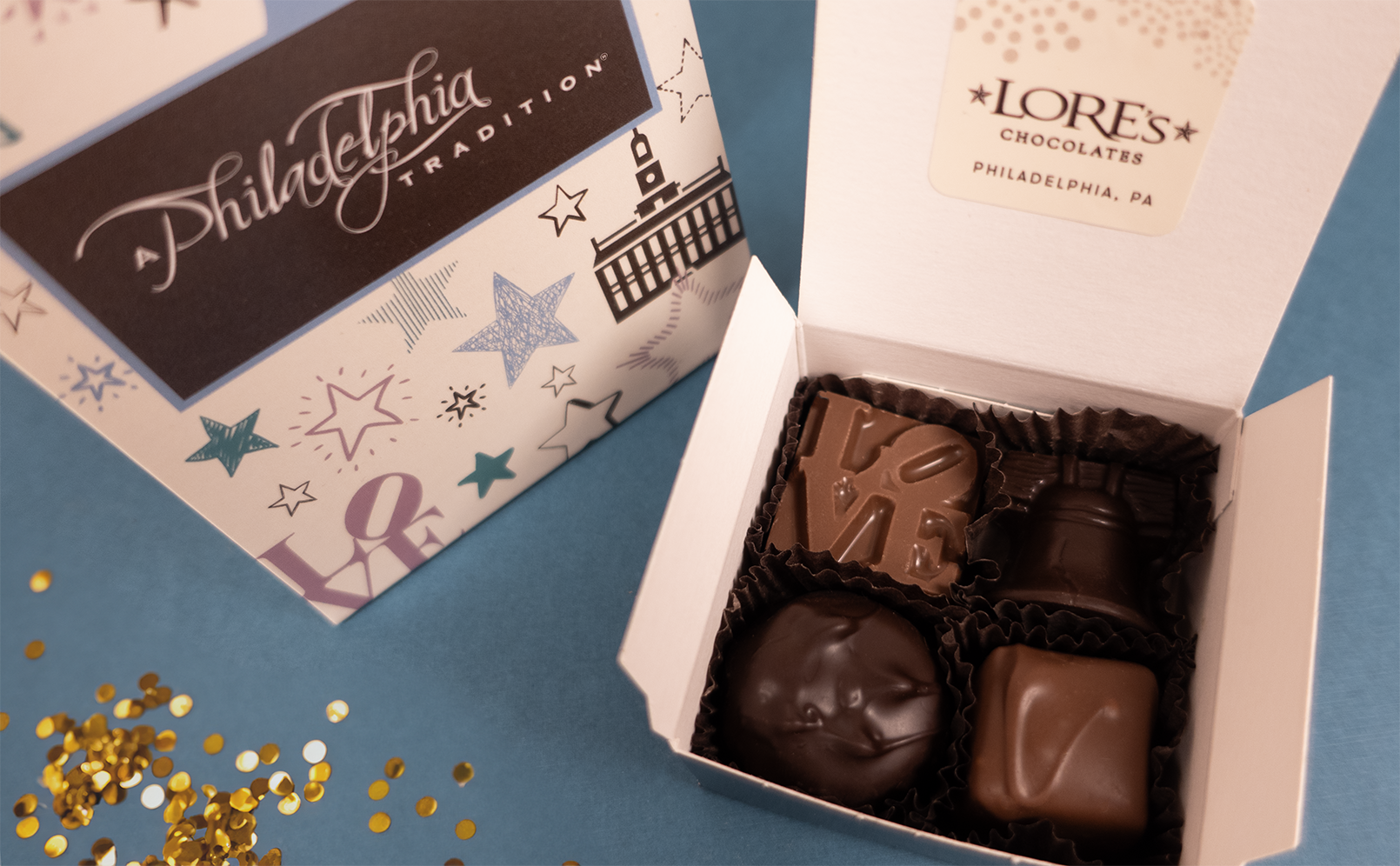 close up of 4 piece chocolate assortment with a philadelphia chocolate liberty bell and LOVE from Lore's Chocolates. blue background with a small pile of gold sequins.