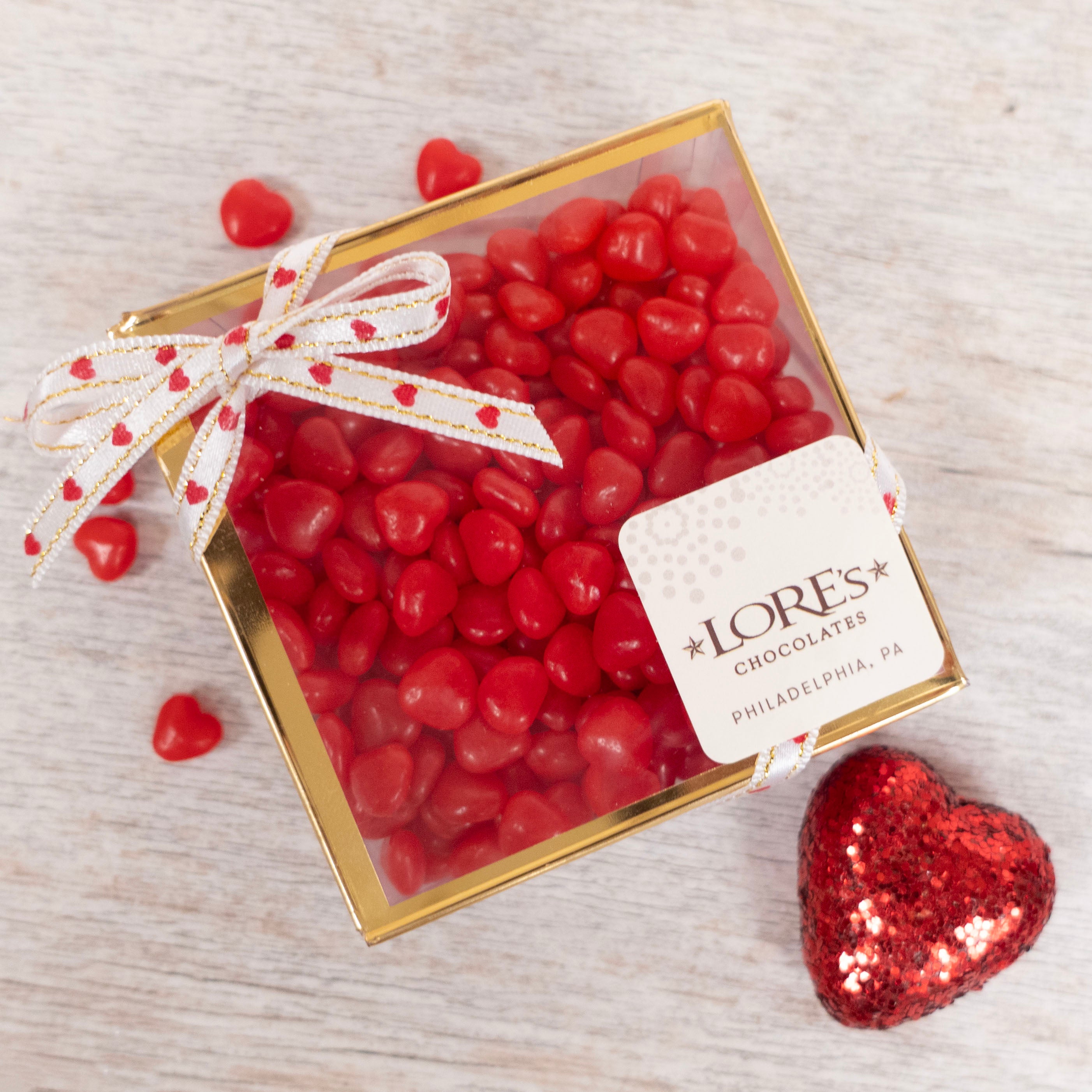 Box of red heart-shaped candies with a bow and Lore's Chocolates label on a light wooden surface.