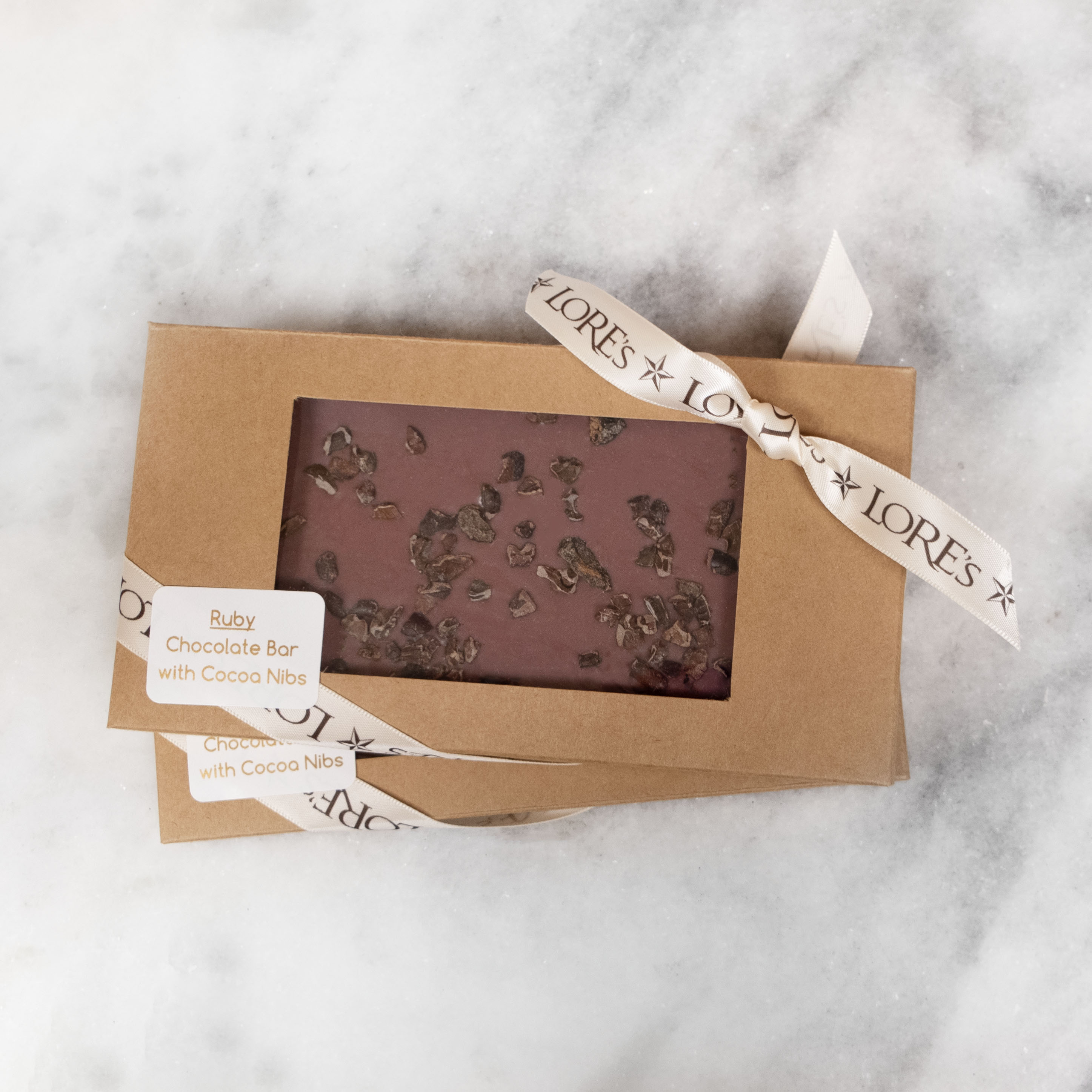 Naturally pink-colored chocolate known as Ruby Chocolate in a Kraft box finished with a Lore's cream-colored Ribbon. Label Reads: Ruby Chocolate Bar with Cocoa Nibs