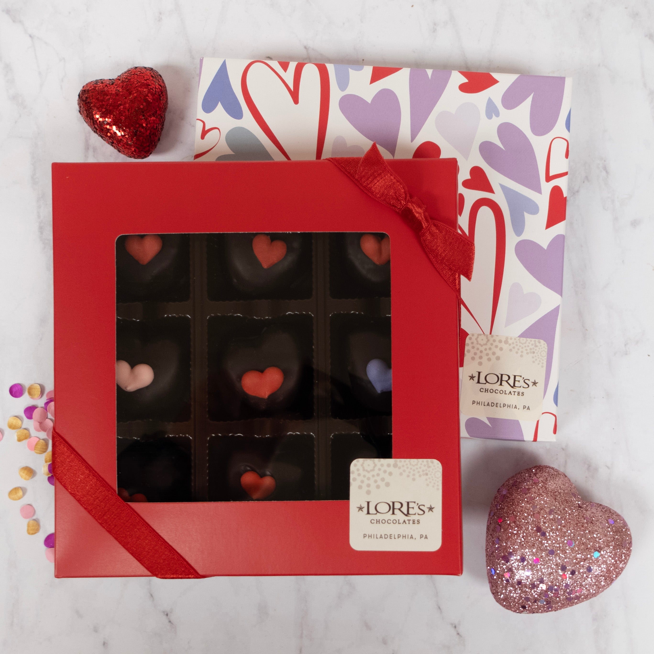 Red box of heart-shaped chocolates with LORES Chocolates branding on a marble surface with decorative hearts and candy.