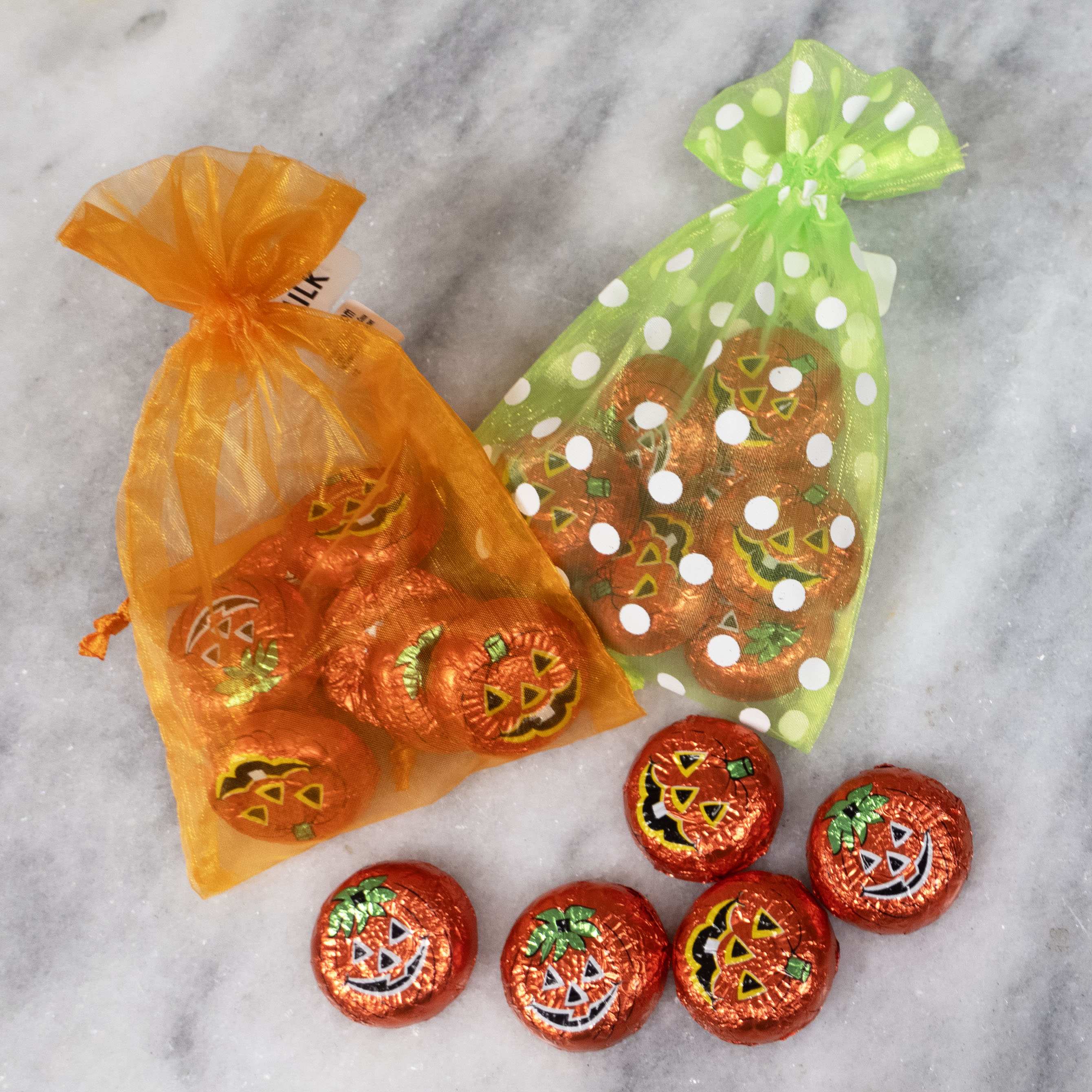 Milk chocolate pumpkin faced candy in orange and green foiled bags, with a few pieces displayed outside the bags on a marble surface.
