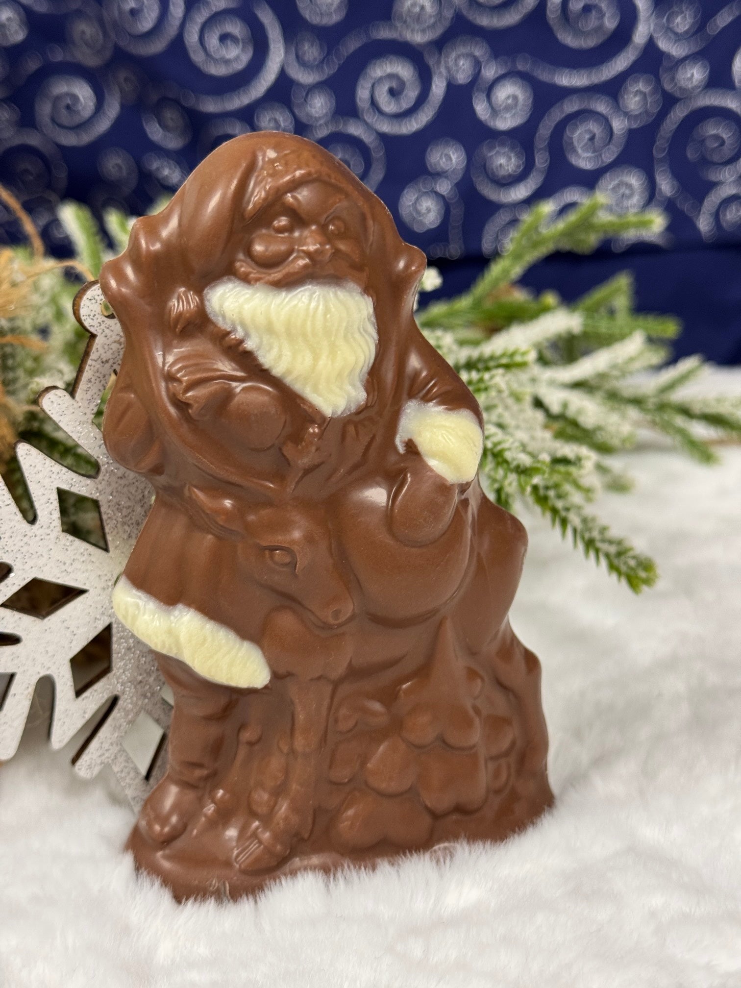 A milk chocolate semi solid figure of Santa Claus, with a white beard, positioned on a white surface with a blue patterned background and decorative snowflakes around.