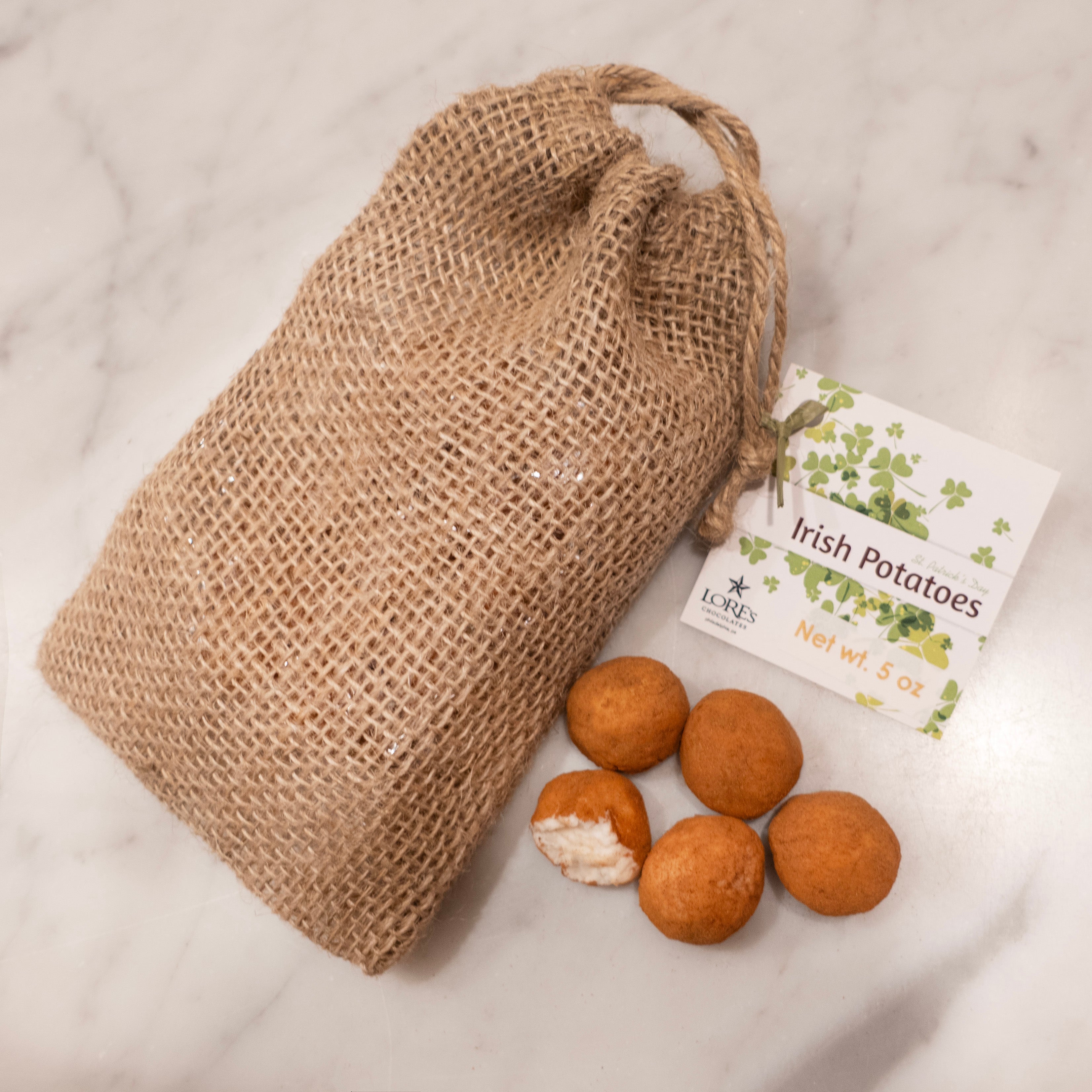 burlap sack from Lore's Chocolates with hang tag reading "Irish Potatoes, Lore's Chocolates, Philadelphia, PA, Net wt 5 oz" Next to the bag are 5 cinnamon covered irish potatoes, one open to show the coconut cream inside