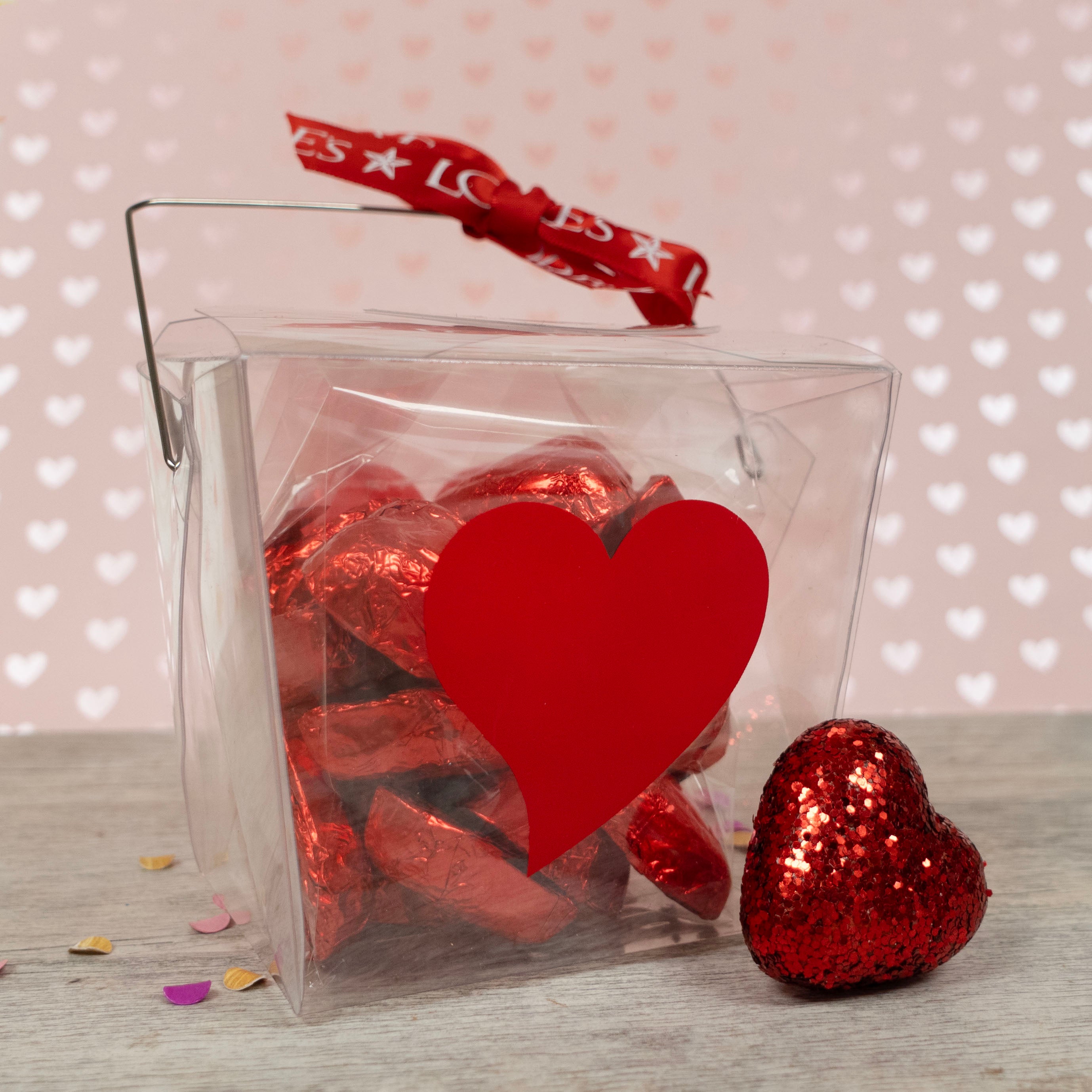 Clear take out box filled with red heart-shaped chocolate wraps on a light wooden surface with a heart-patterned background.
