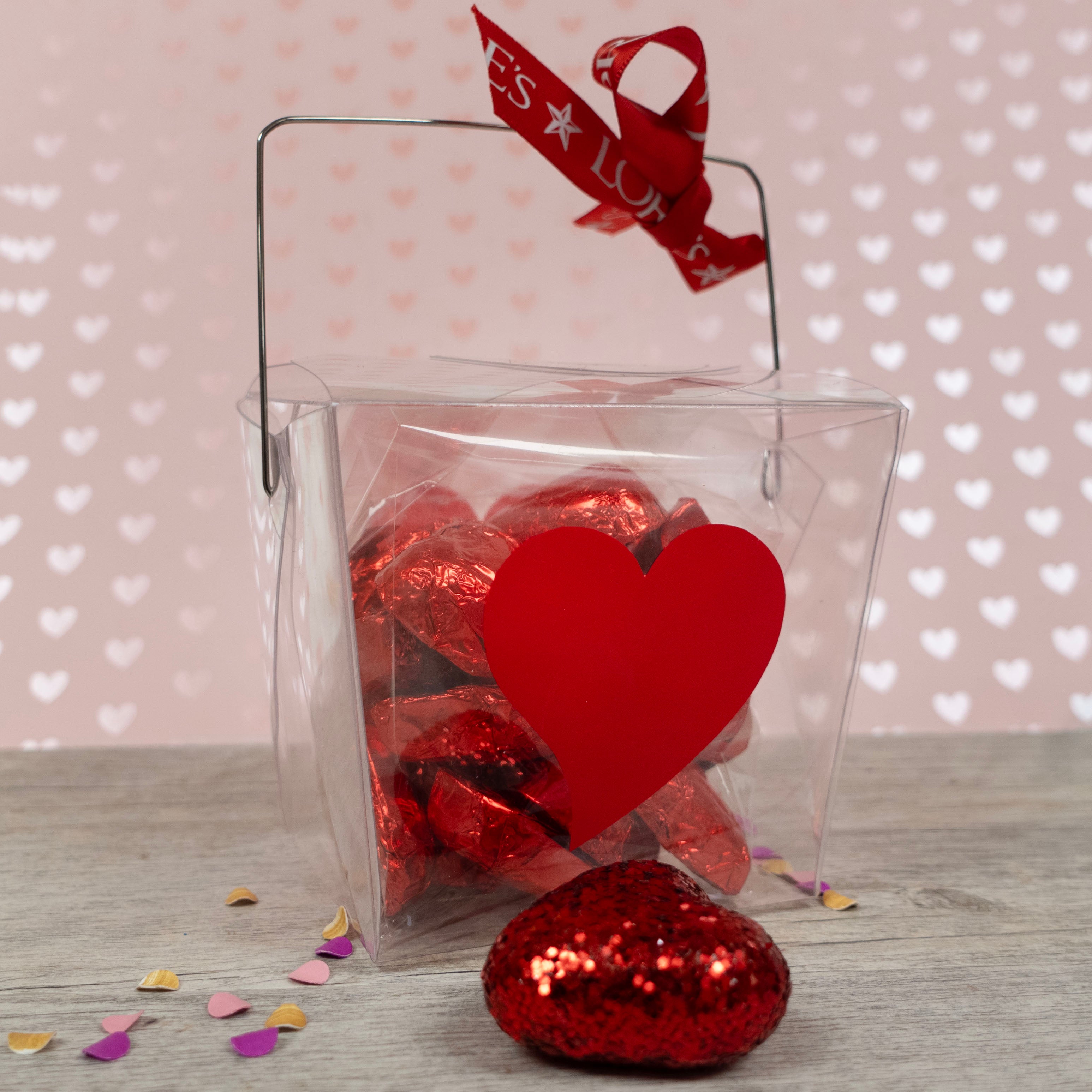 Clear takeout-style gift box with red heart-shaped tag and foil-wrapped candies on a heart-patterned background