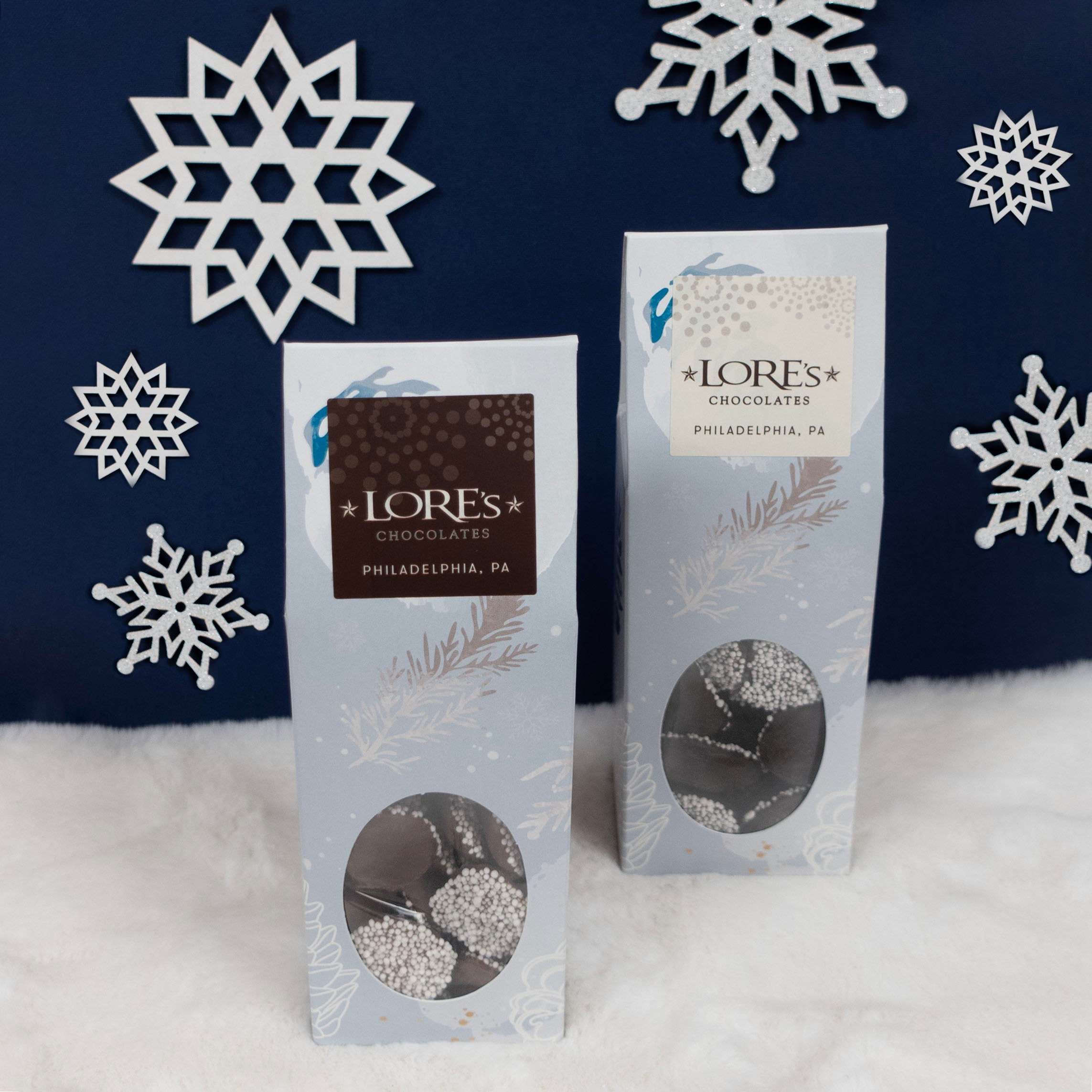 Two packages of Lores Chocolates nonpareils with decorative snowflakes in the background