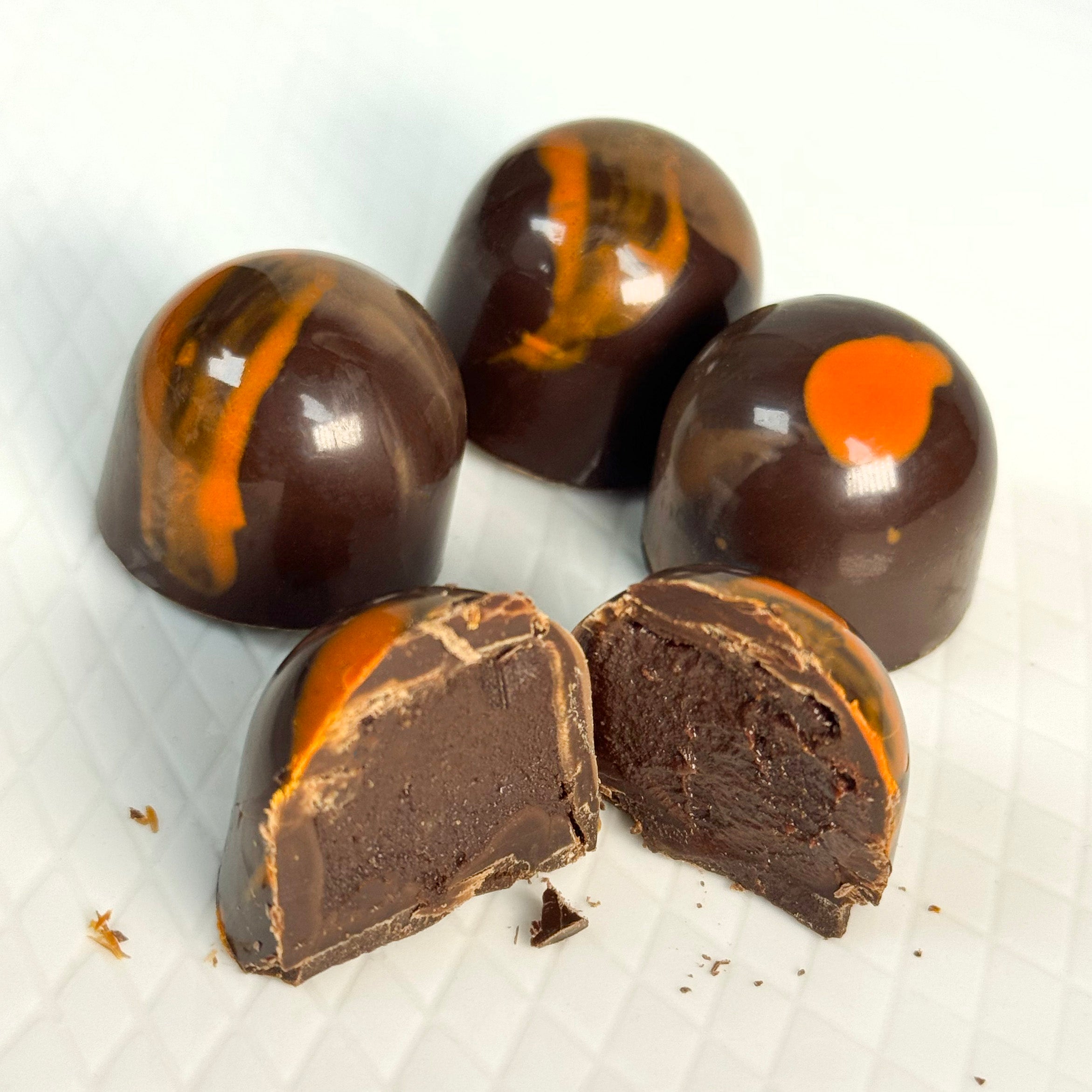 Four glossy dark chocolate truffles with orange swirls and one cut open to show the spiced dark chocolate ganache filling, arranged on a white textured surface.