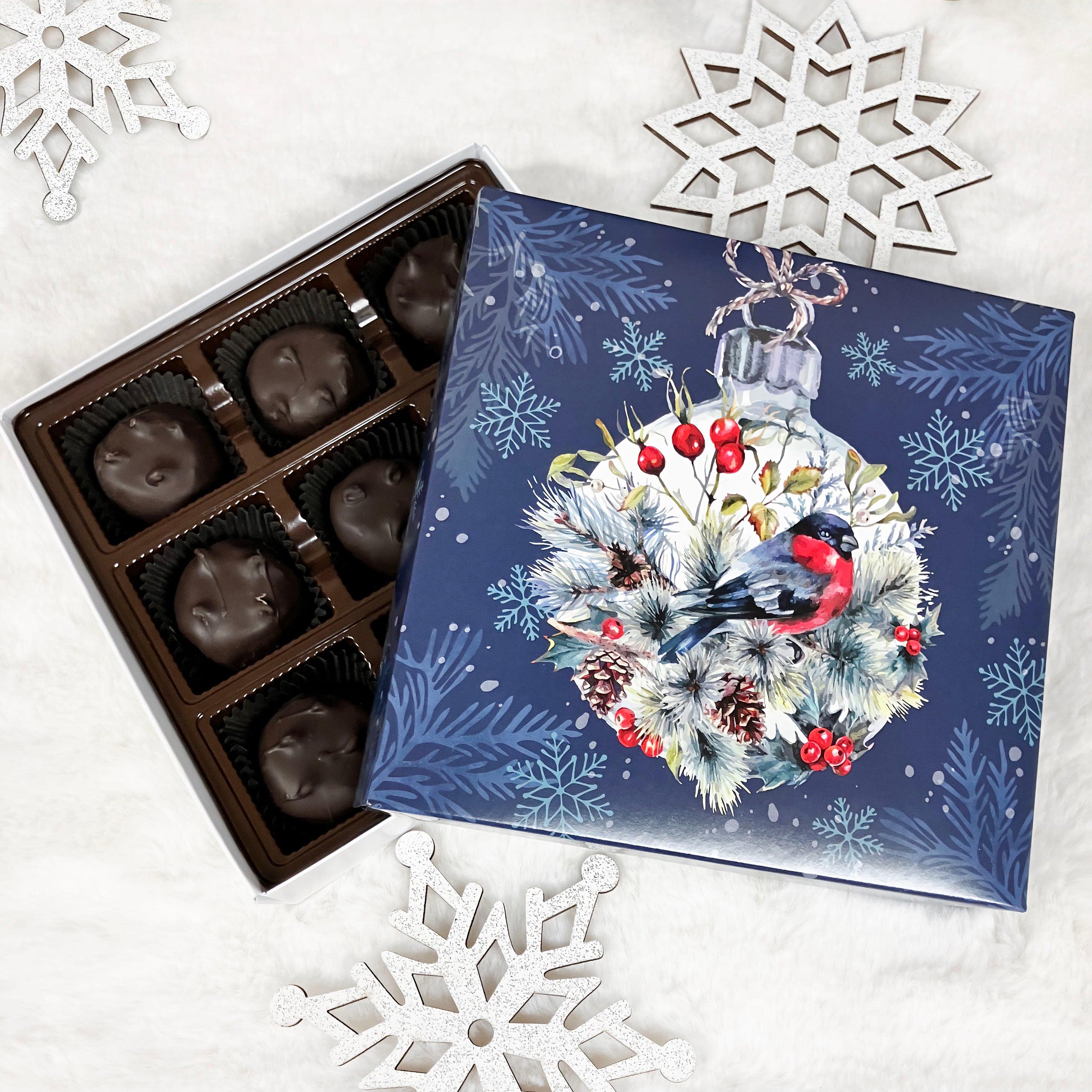 Box of chocolates with a decorative cover featuring a bird and Christmas elements on a white surface with snowflake decorations.