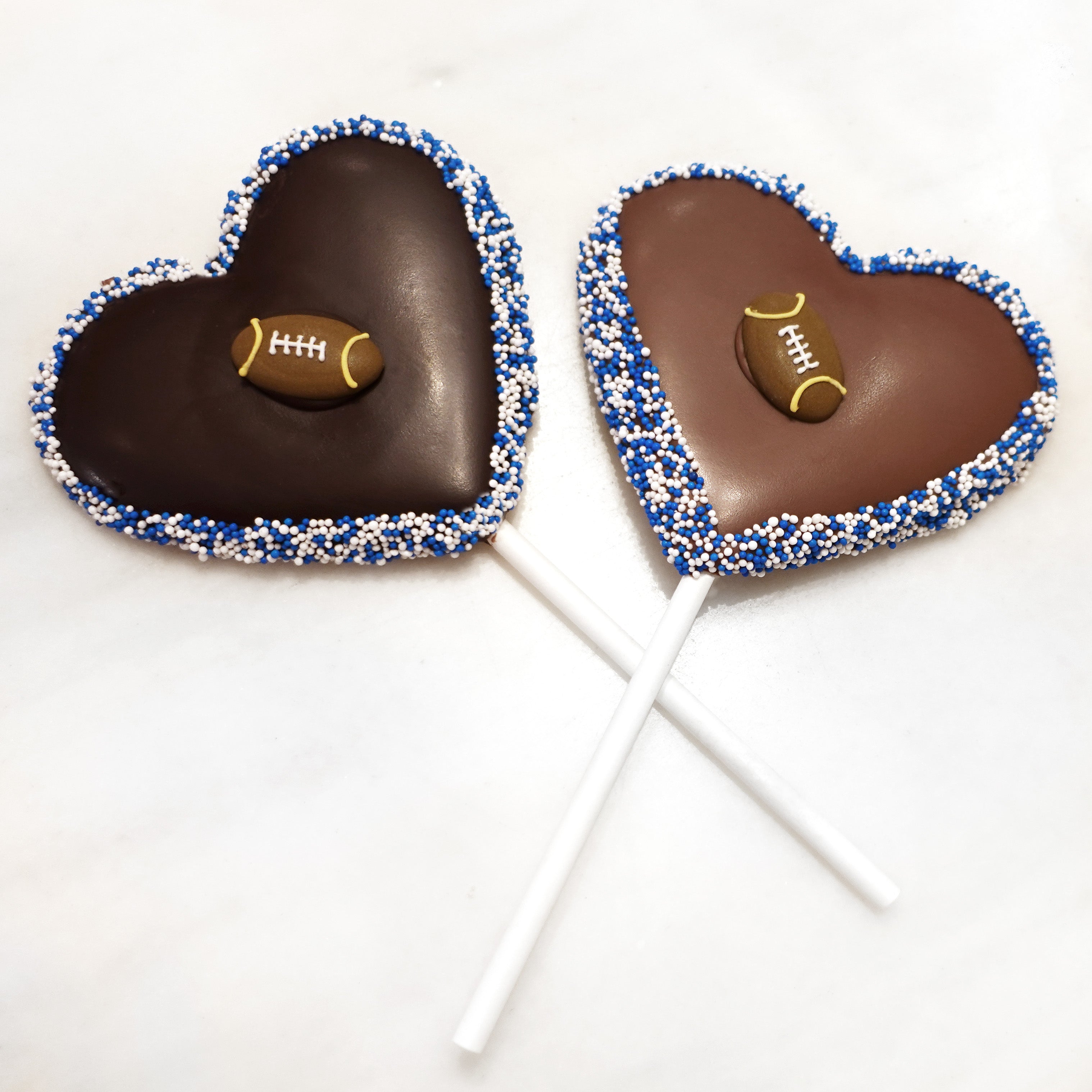 dark chocolate (left) and milk chocolate (right) heart shaped lollipops with blue and white nonpareils seeds on edge and a sugar football in the center - made and sold by lore's chocolates