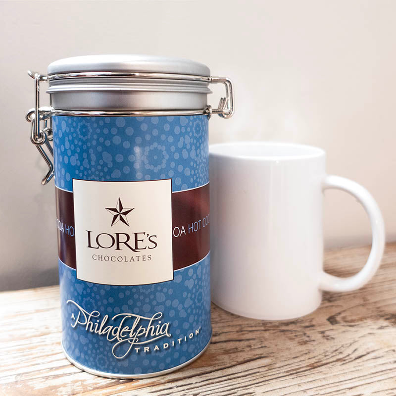 Blue cylindrical container of Hot Cocoa Mix with a white label "Lore's Chocolates" and "A Philadelphia Tradition" next to a white steaming mug on a wooden surface