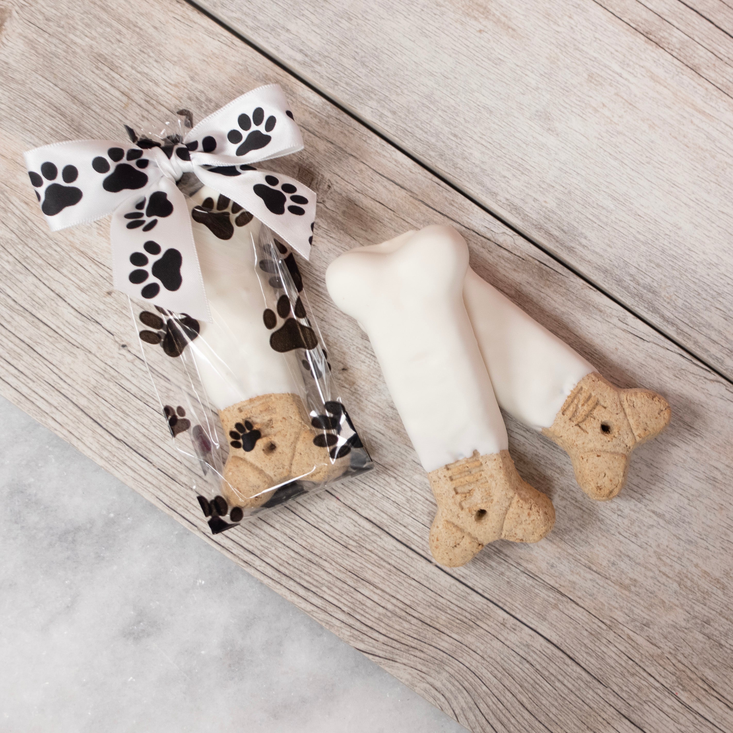 Two Milk Bone Dog biscuits dipped in Lore's Chocolates (dog safe) white yogurt - left dog treat is packaged in a paw print cellophane bag with black and white paw prints bow on top