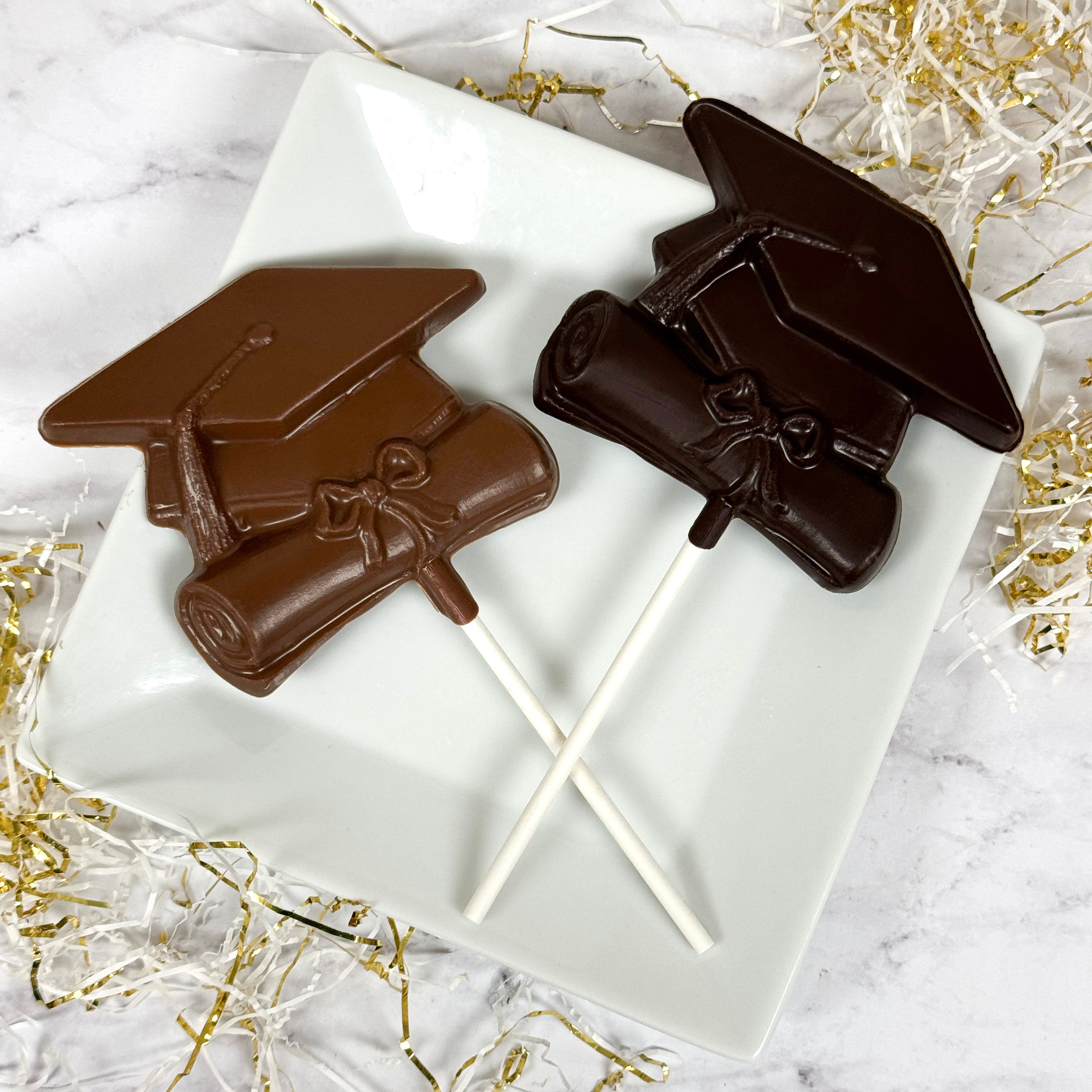 Chocolate graduation caps on lollipop sticks with a white plate on a marble surface.