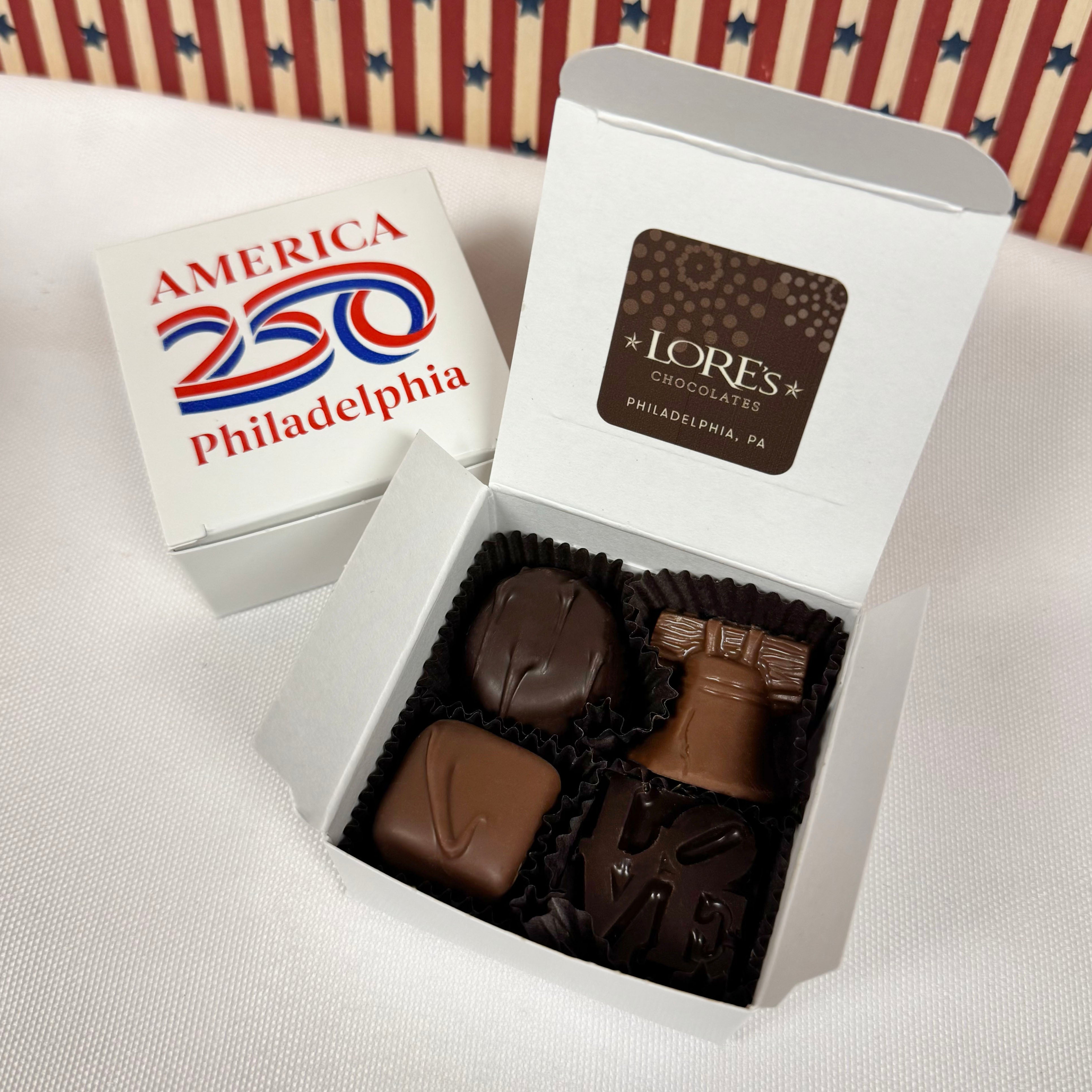 4 piece gift box of chocolates with a 'Lore's Chocolates' label, featuring the America 250 Philadelphia logo in the background. Milk Chololate Liberty Bell, Dark Philly's LOVE statue, milk chocolate caramel and a dark butter cream.