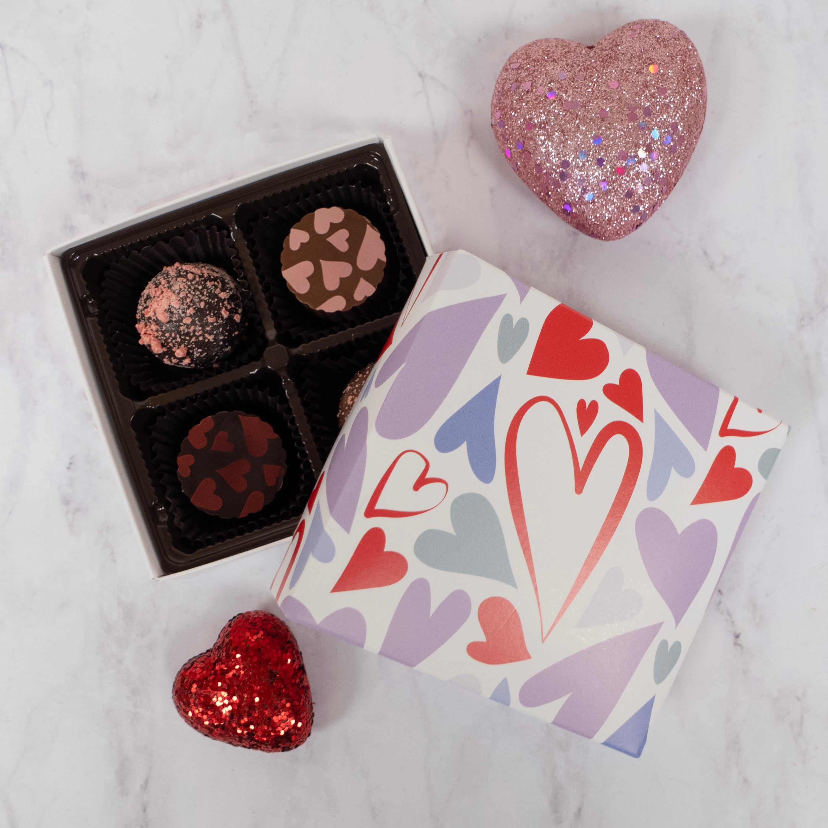 Box of chocolates with a heart-shaped glittery object and box has heart designs on a marble surface.