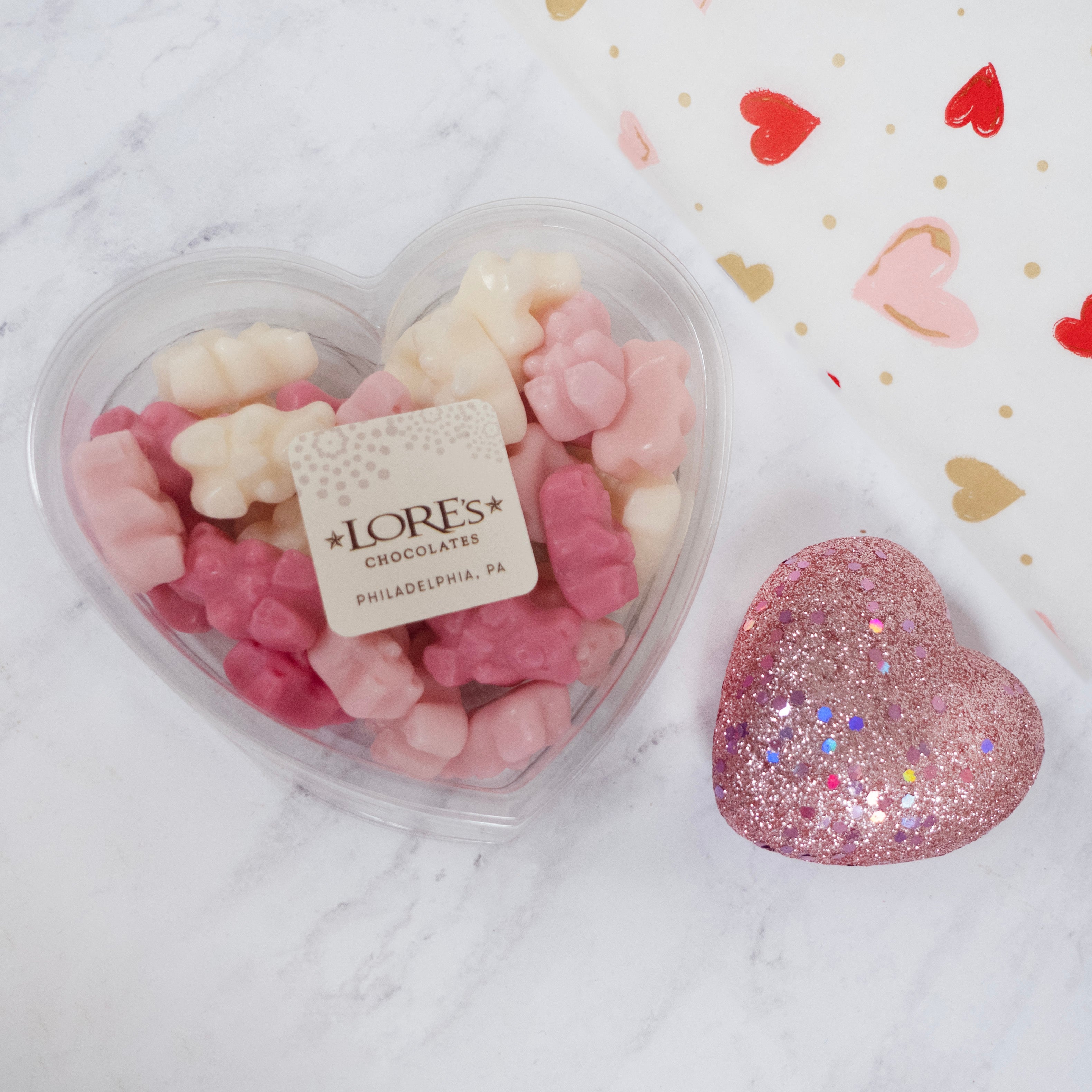 Heart-shaped container of pink and white gummy bears with a glittery pink heart next to it on a marble surface.