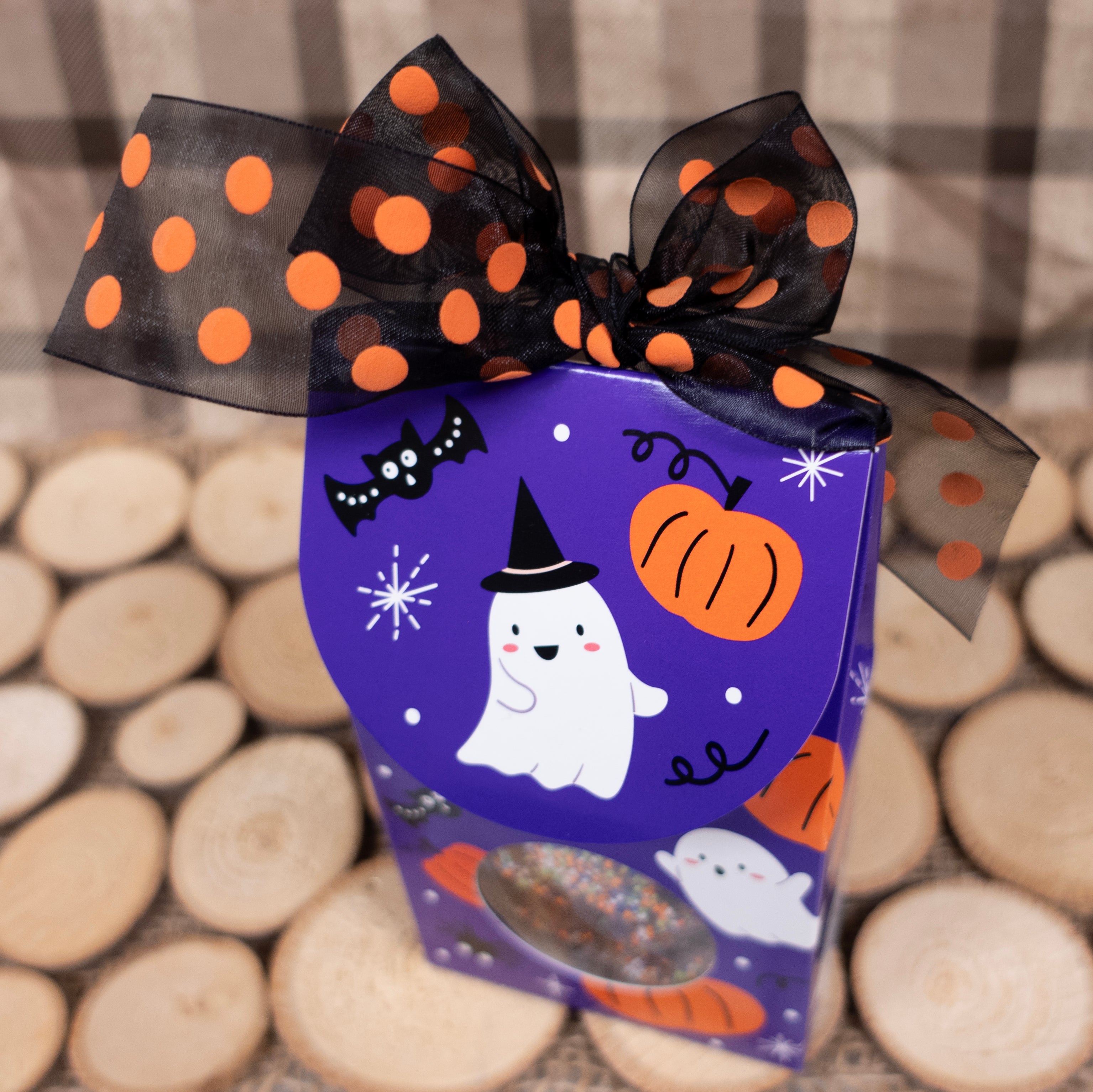 Halloween-themed gift box with a purple lid featuring ghost and pumpkin illustrations, tied with a black and orange polka dot ribbon.