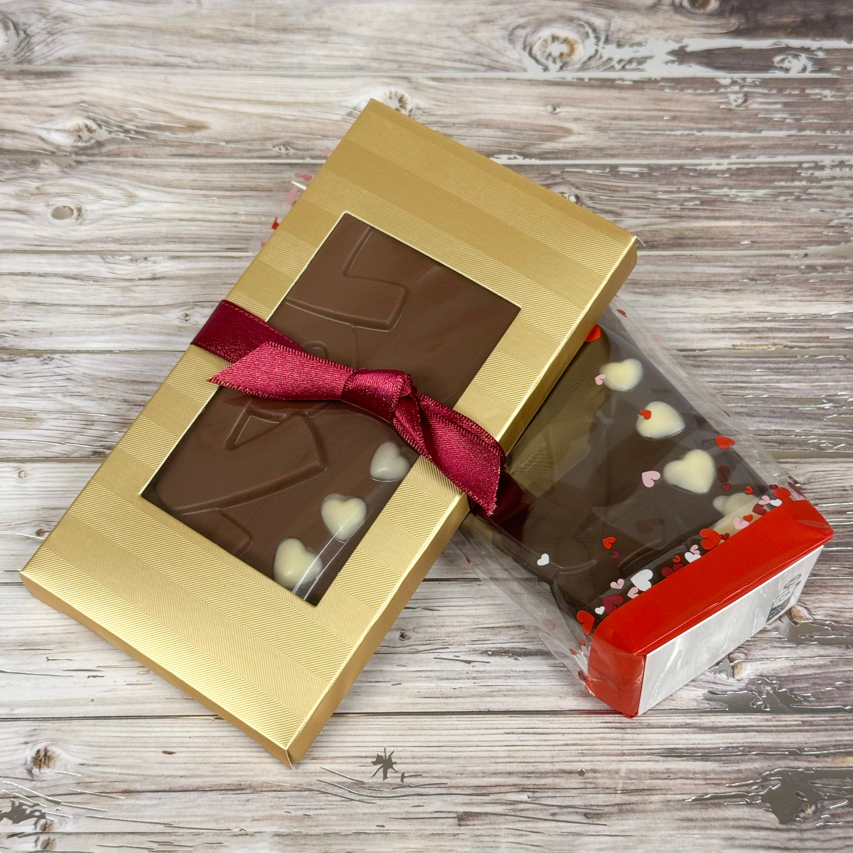 Chocolate bar with LOVE and white chocoalte hearts in a gold box with a red ribbon on a wooden surface. made by Lore's Chocolates in Philadelphia