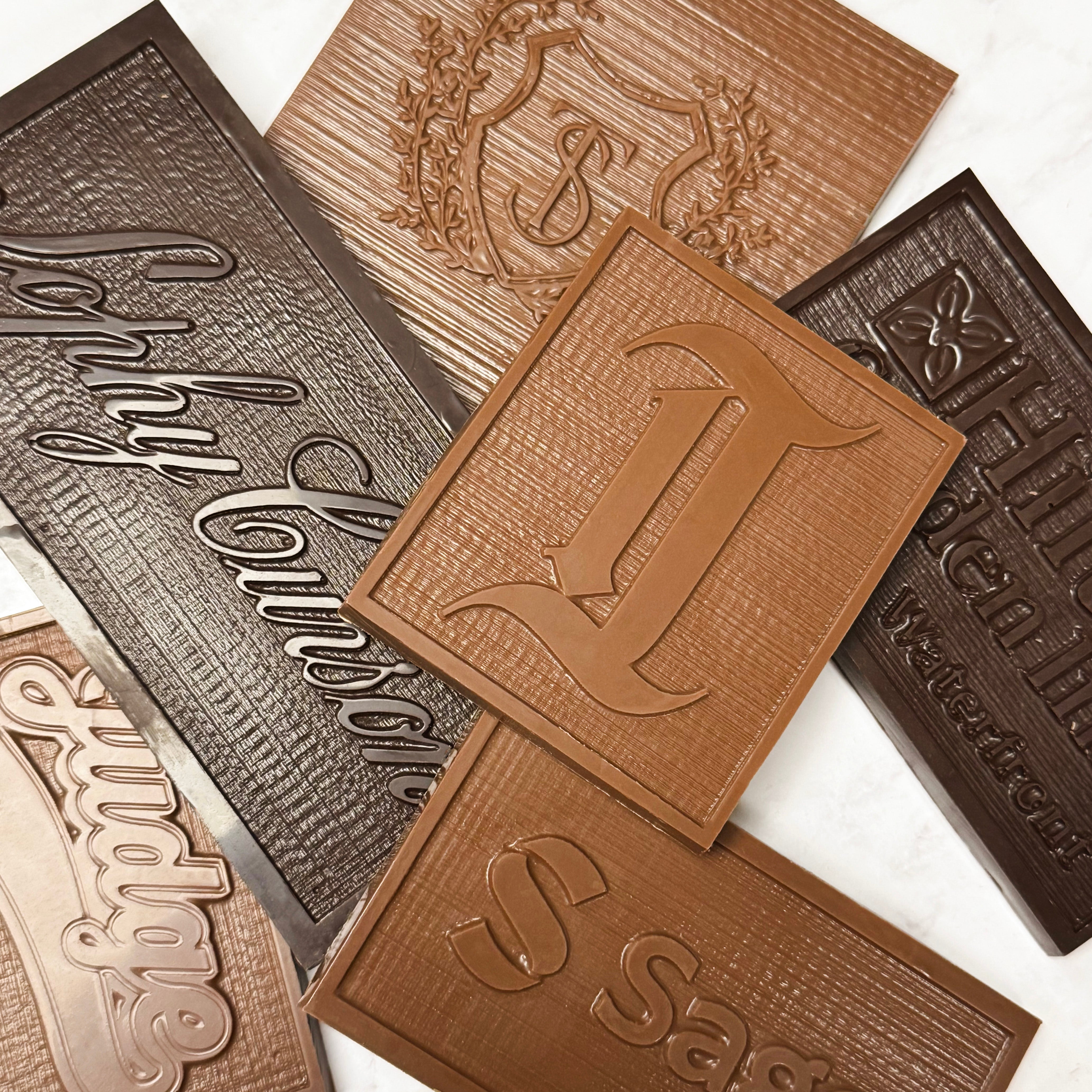 A set of custom embossed chocolate blocks in various shades of brown, featuring monogrammed initials, business logos, and decorative crest designs, (most prominent being the Philadelphia Inquirer), arranged on a light marble background. - handmade by Lore's Chocolates in Philadelphia