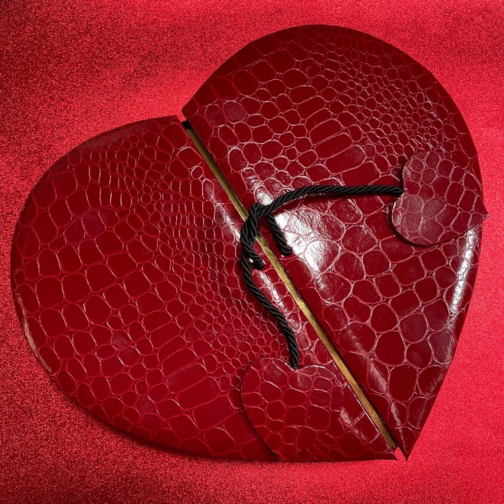 Heart shaped box with a crocodile/alligator leather pattern, opens in the middle with black cord with small alligator heart on either door.  Background is a sparkly red (Box Contains Artisan Truffles from Lore's Chocolates)