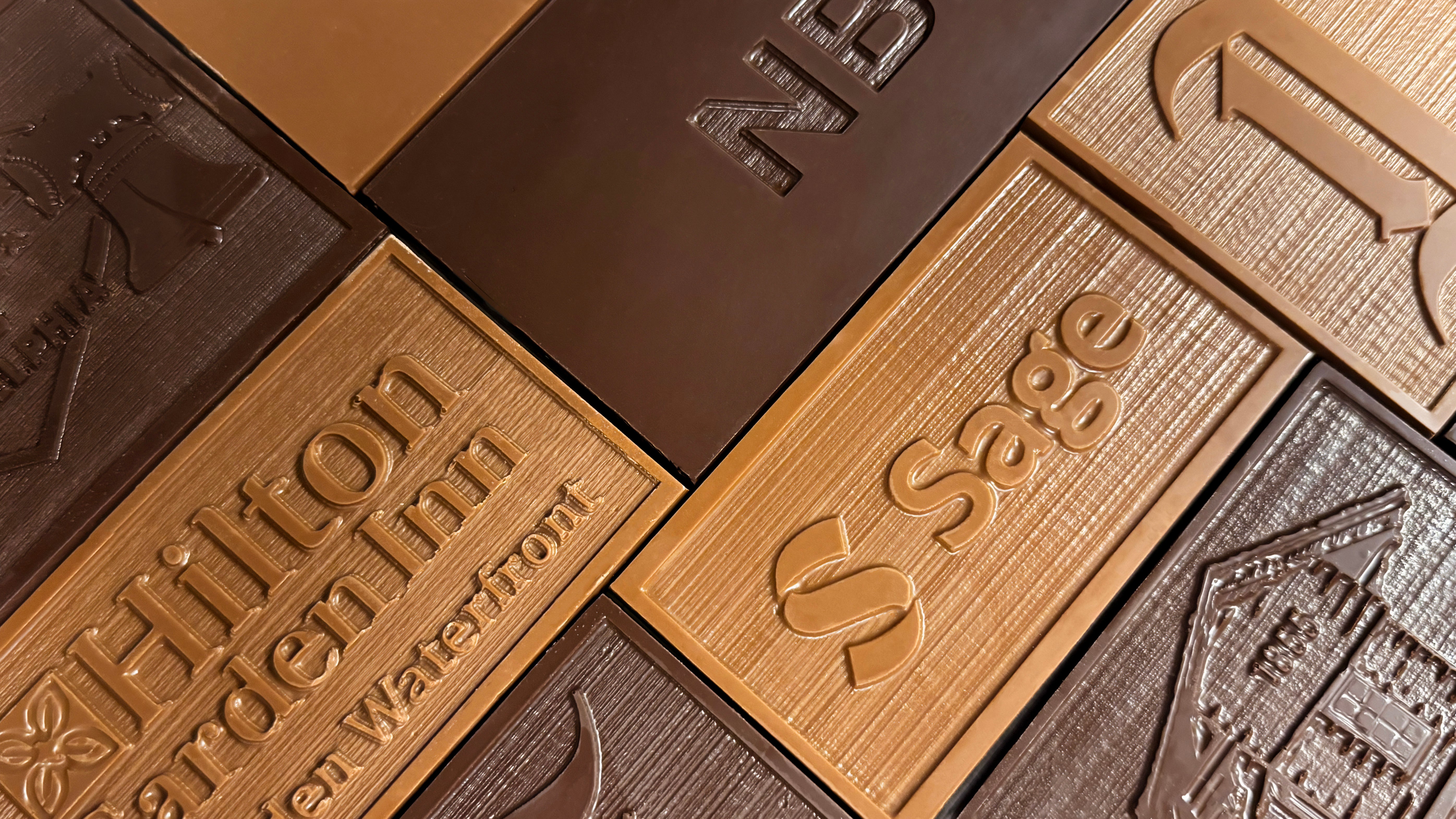 A close-up arrangement of branded chocolate bars in multiple shades of brown, including embossed text such as 'Hilton Garden Inn' and 'Sage'.