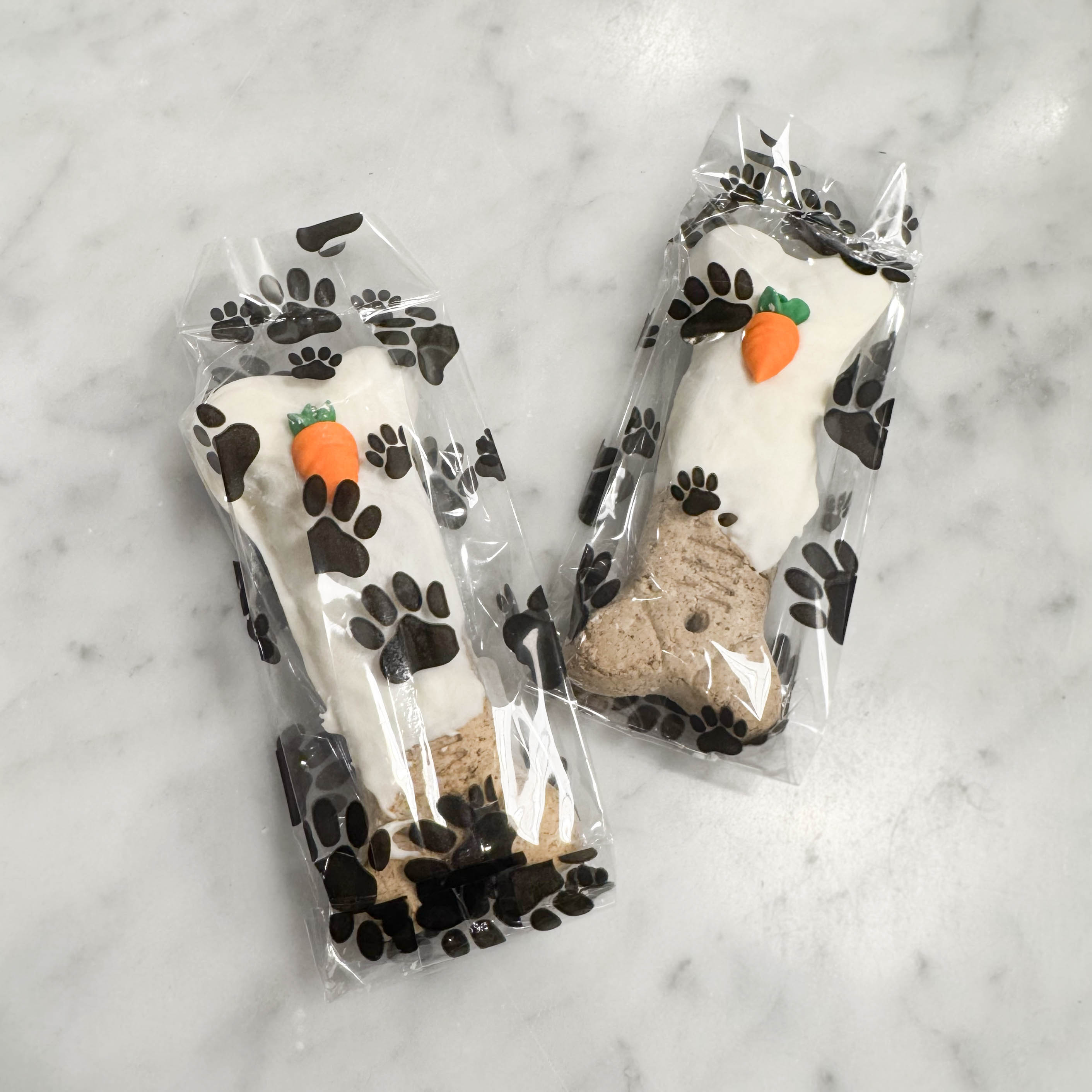 Two packages of dog treats shaped like bones with carrot candies in a paw print bag on a marble surface.