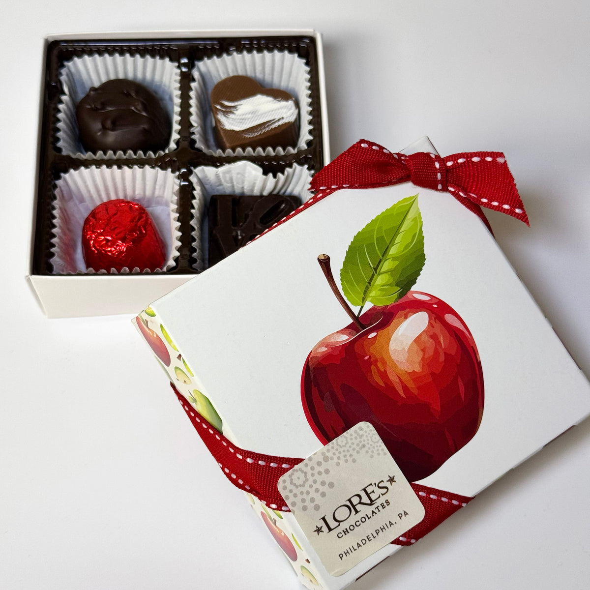 A boxed assortment of chocolates with one box open to show four chocolates inside white paper cups, and a second white gift box in front with a red ribbon and an apple illustration. The front box has a visible tag reading LORE'S Chocolates Philadelphia PA