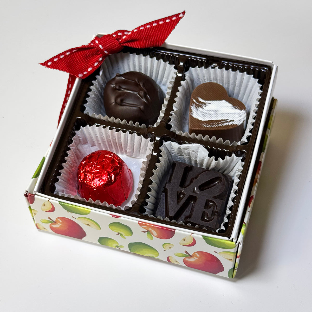 A boxed assortment of four chocolates in a white gift box with a red ribbon bow. The chocolates include a dark chocolate vanilla buttercream, a milk chocolate caramel-filled heart with white drizzle, a red foil-wrapped milk chocolate, and a dark chocolate square embossed with the word 'LOVE'. Made in Philadelphia