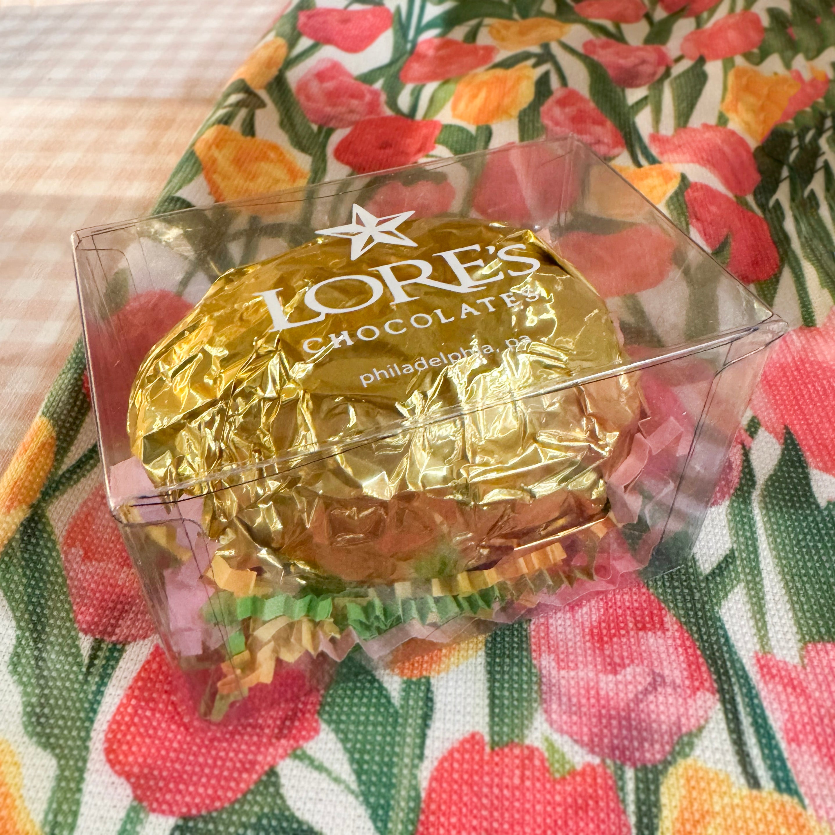 Gold-wrapped chocolate egg in a clear box on a floral-patterned surface. box says "lore's chocolates philadelphia, pa"