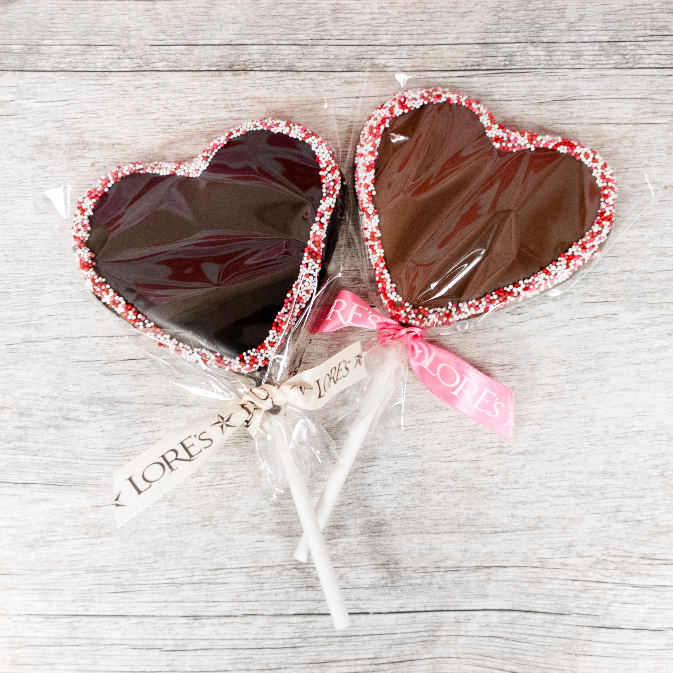 two heart shaped chocolate lollipops outlined in white, pink, and red nonpareil seeds with Lore's Chocolates ribbons - dark chocolate on left, milk chocolate on right