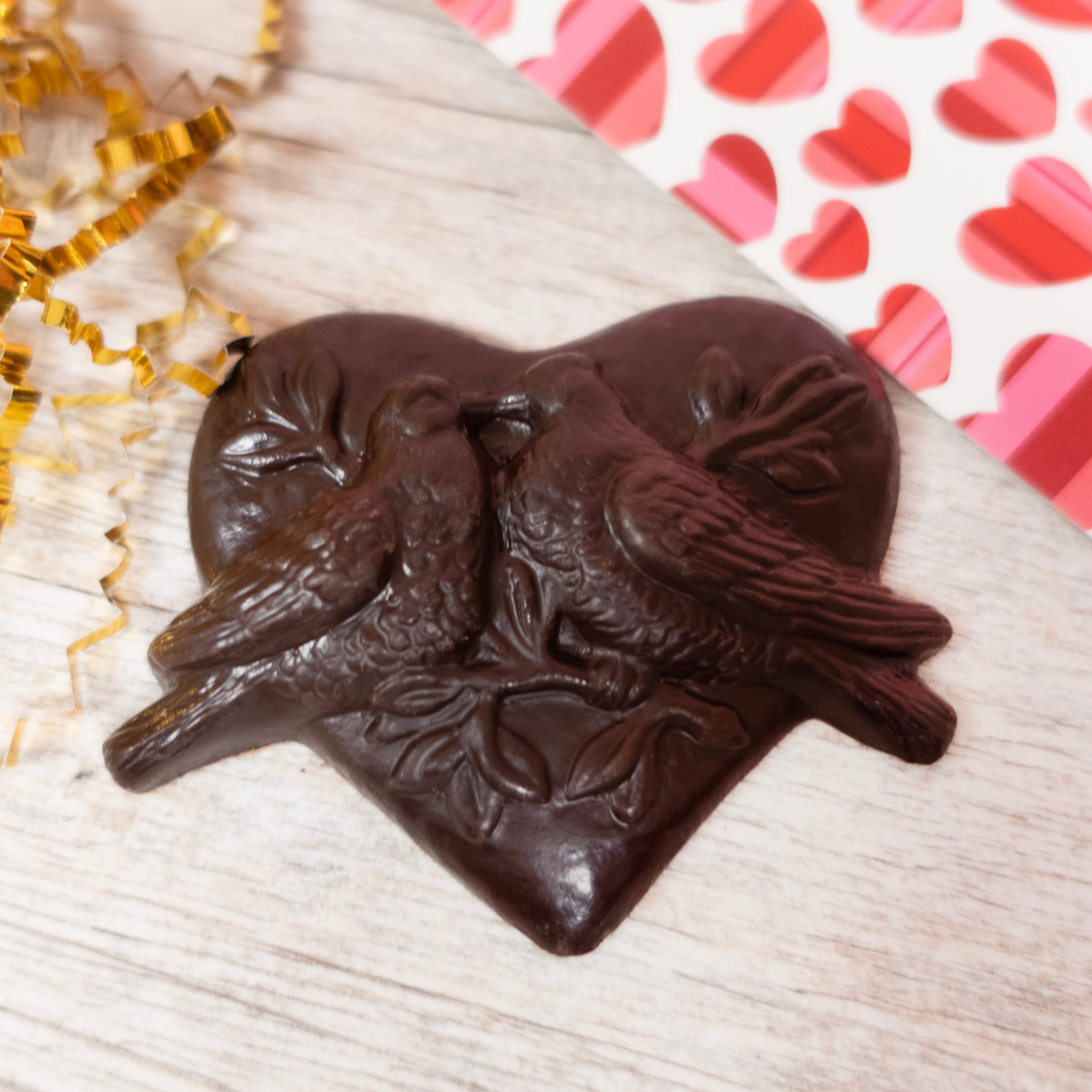 solid chocolate mold: lovebirds kissing on a branch, surrounded by a heart in Lore's Dark chocolate. Hearts and gold foiled crinkle paper in background.