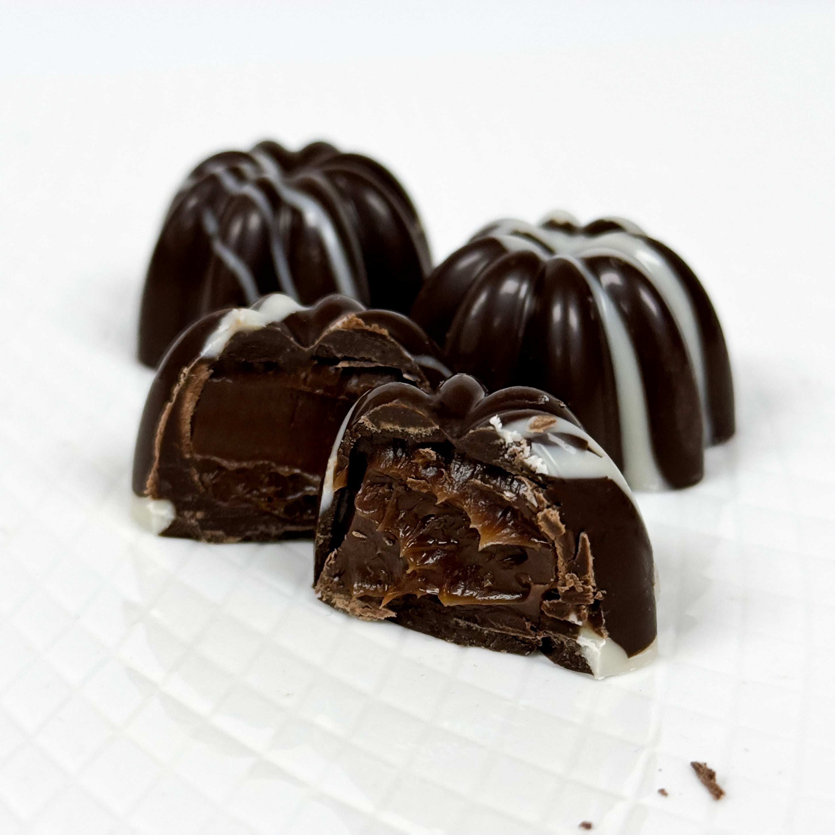 Three chocolate ganache truffles on a white textured surface, with one cut open to show a dark chocolate filling and white drizzle on top.
