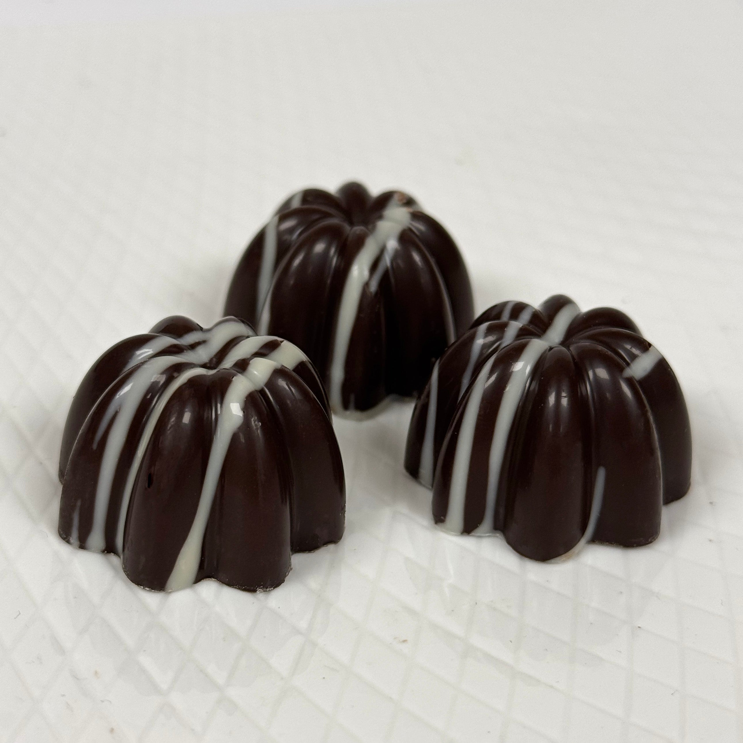 Three small dark chocolate confections with white drizzle, arranged on a white textured surface.