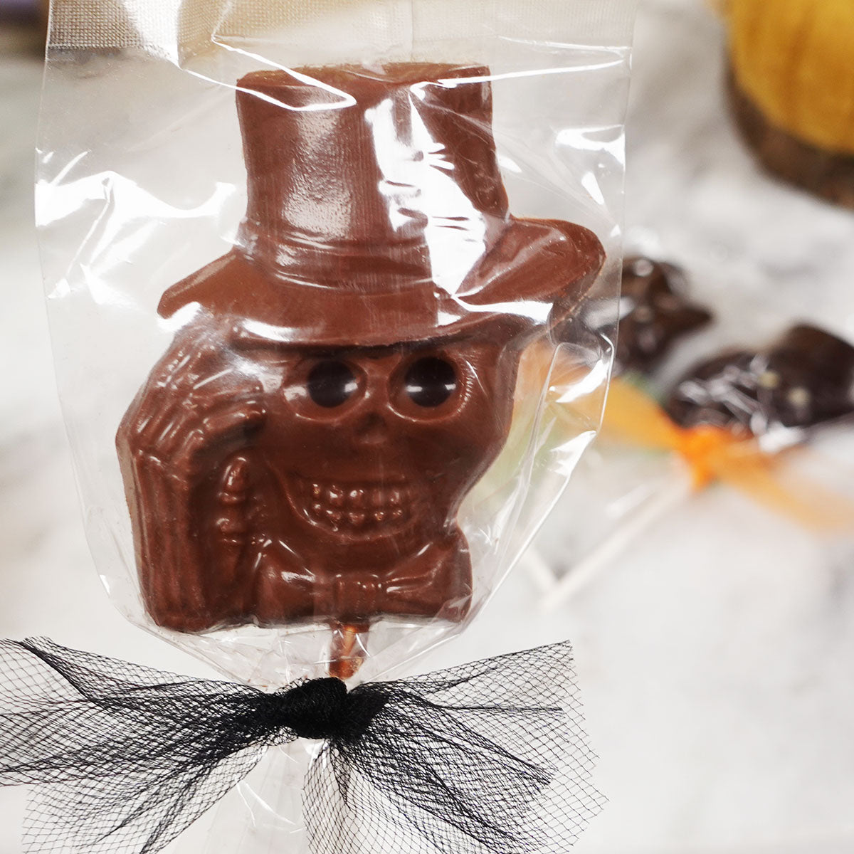 A chocolate lollipop with a skeleton character design, wrapped in a clear plastic wrapper with a black bow on the bottom, displayed on a marble surface.